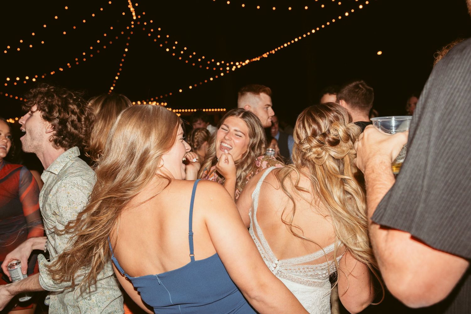 Party guests laughing and dancing together under string lights at an evening celebration.