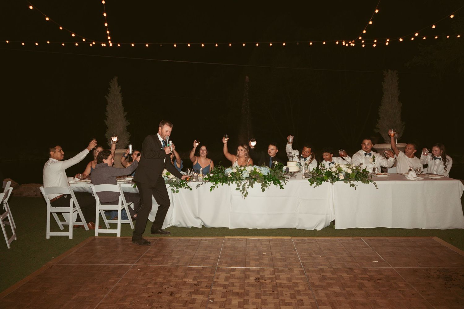 Long reception table decorated with greenery and string lights overhead at outdoor wedding venue.