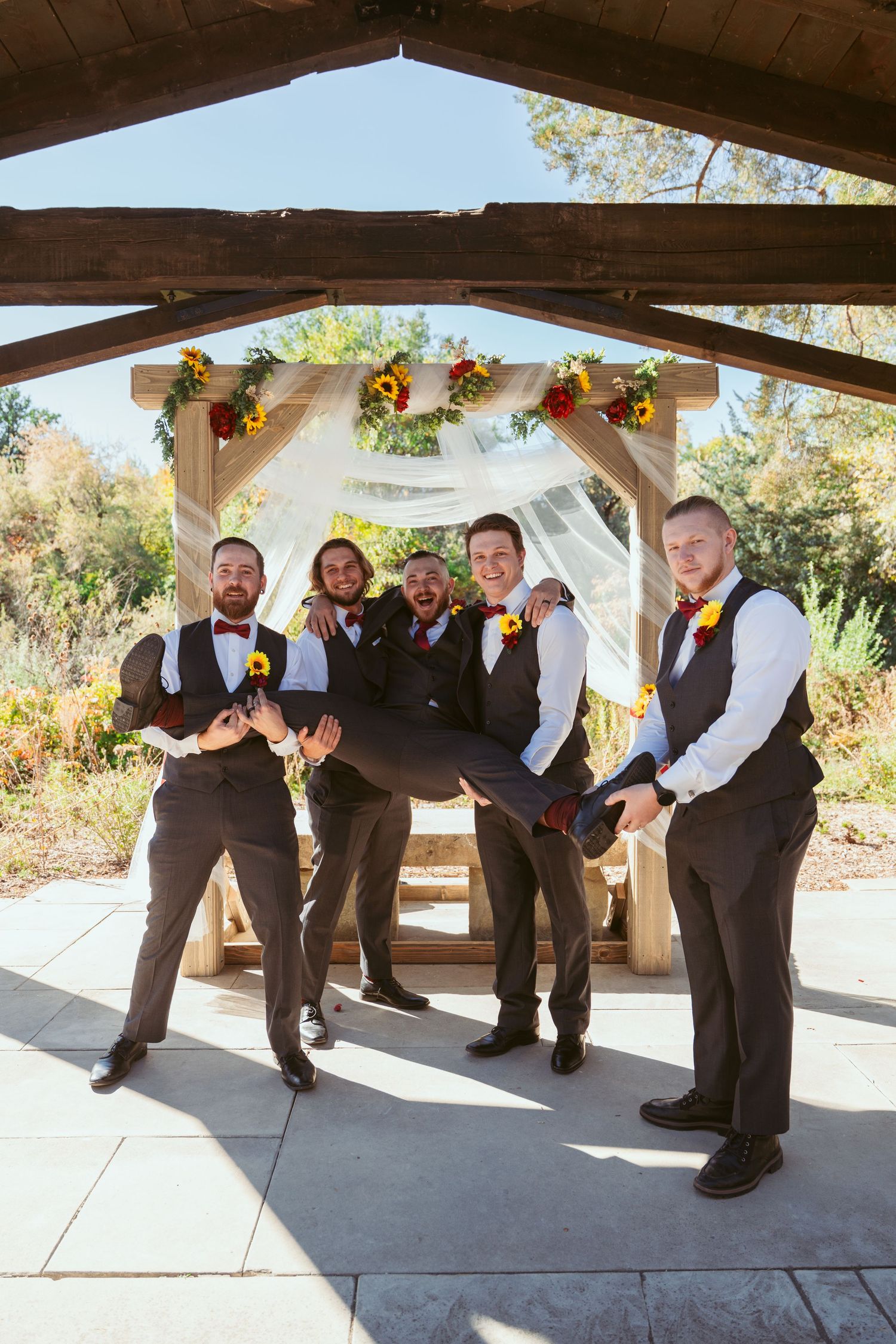 Wedding party members pose playfully in a line under wooden pavilion with fall decorations.