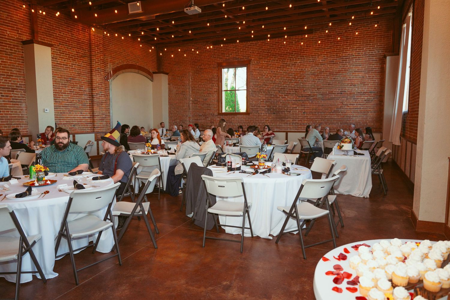 Wedding reception venue with exposed brick walls, string lights, and round tables set for dining.