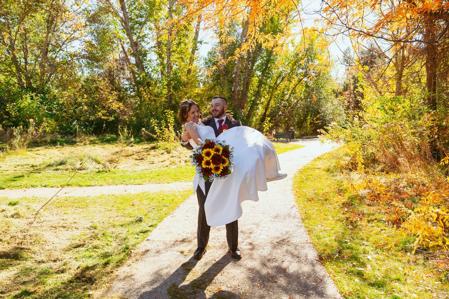 A romantic autumn wedding photo of a couple on a sunlit path surrounded by golden fall foliage.
