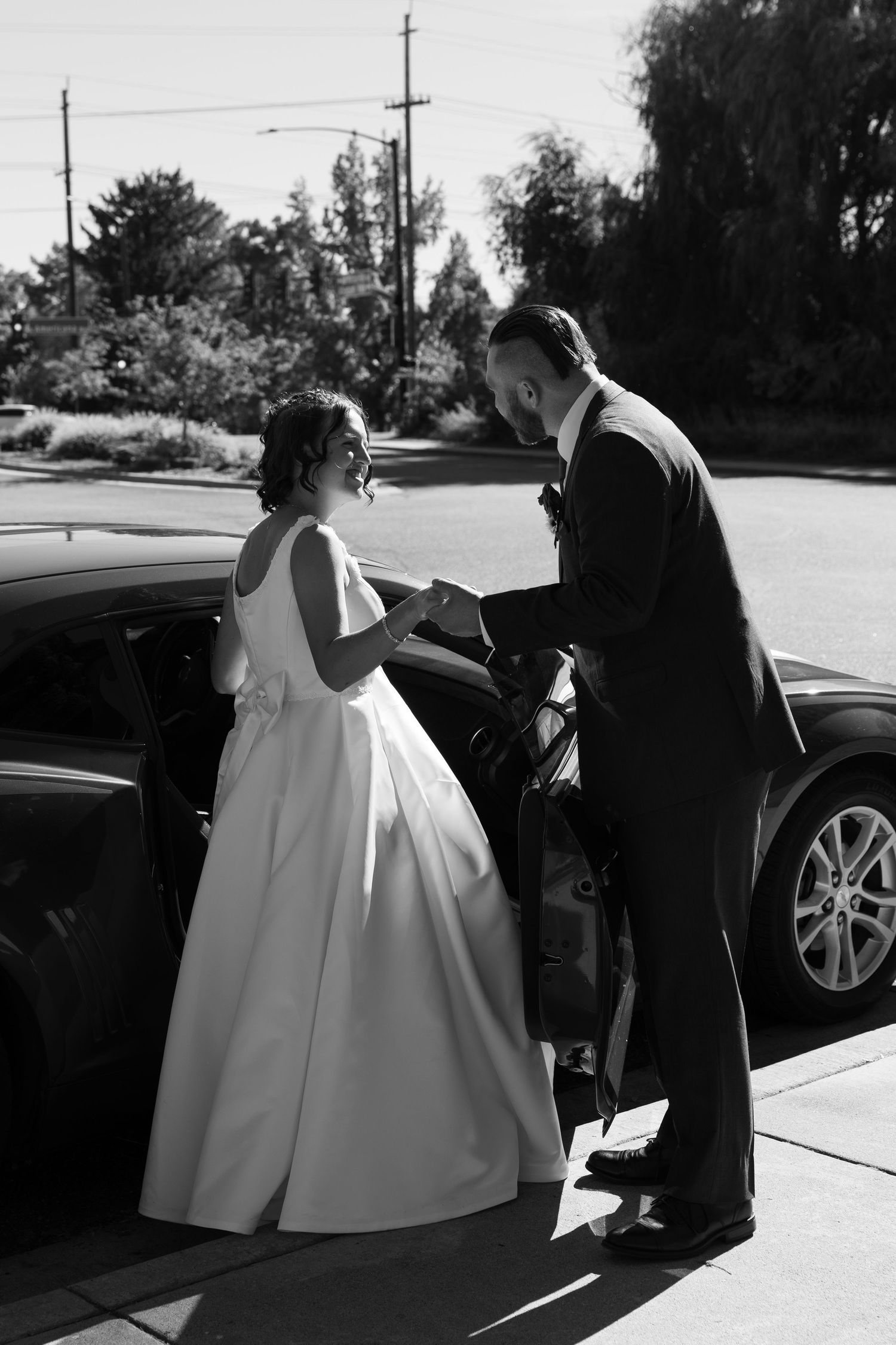 Black and white series showing couple in formal attire near luxury car on wedding day.