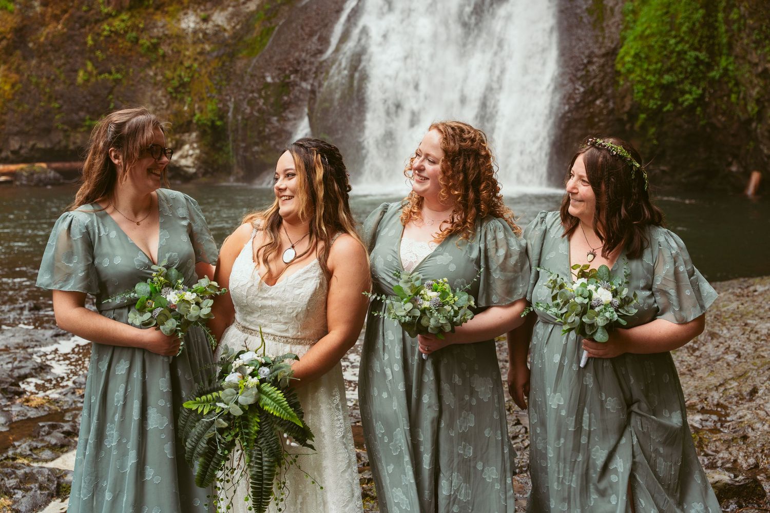 Wedding party laughing together while holding bouquets near a scenic forest waterfall.