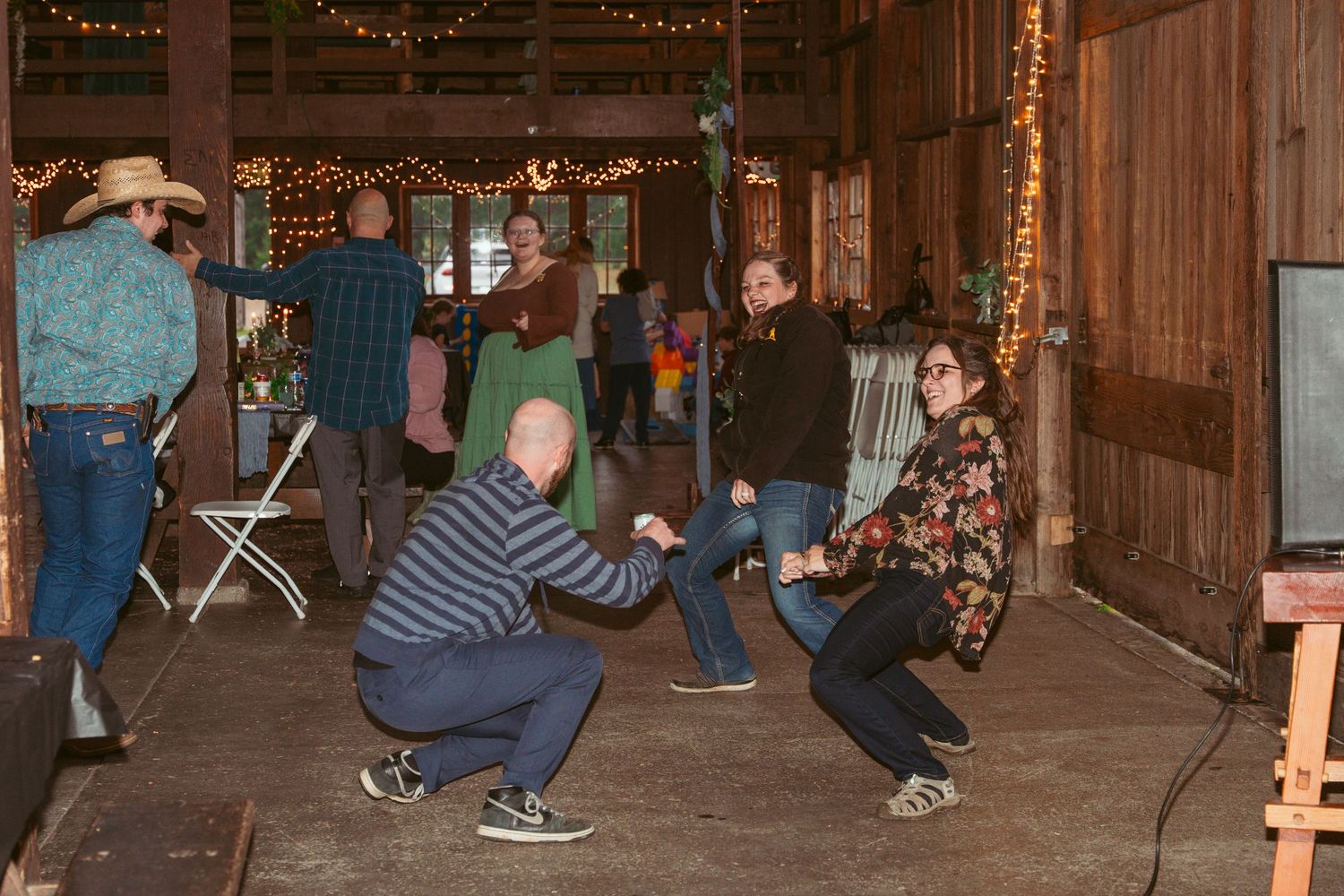 People dancing in lines on a wooden floor under twinkling lights at a barn event.
