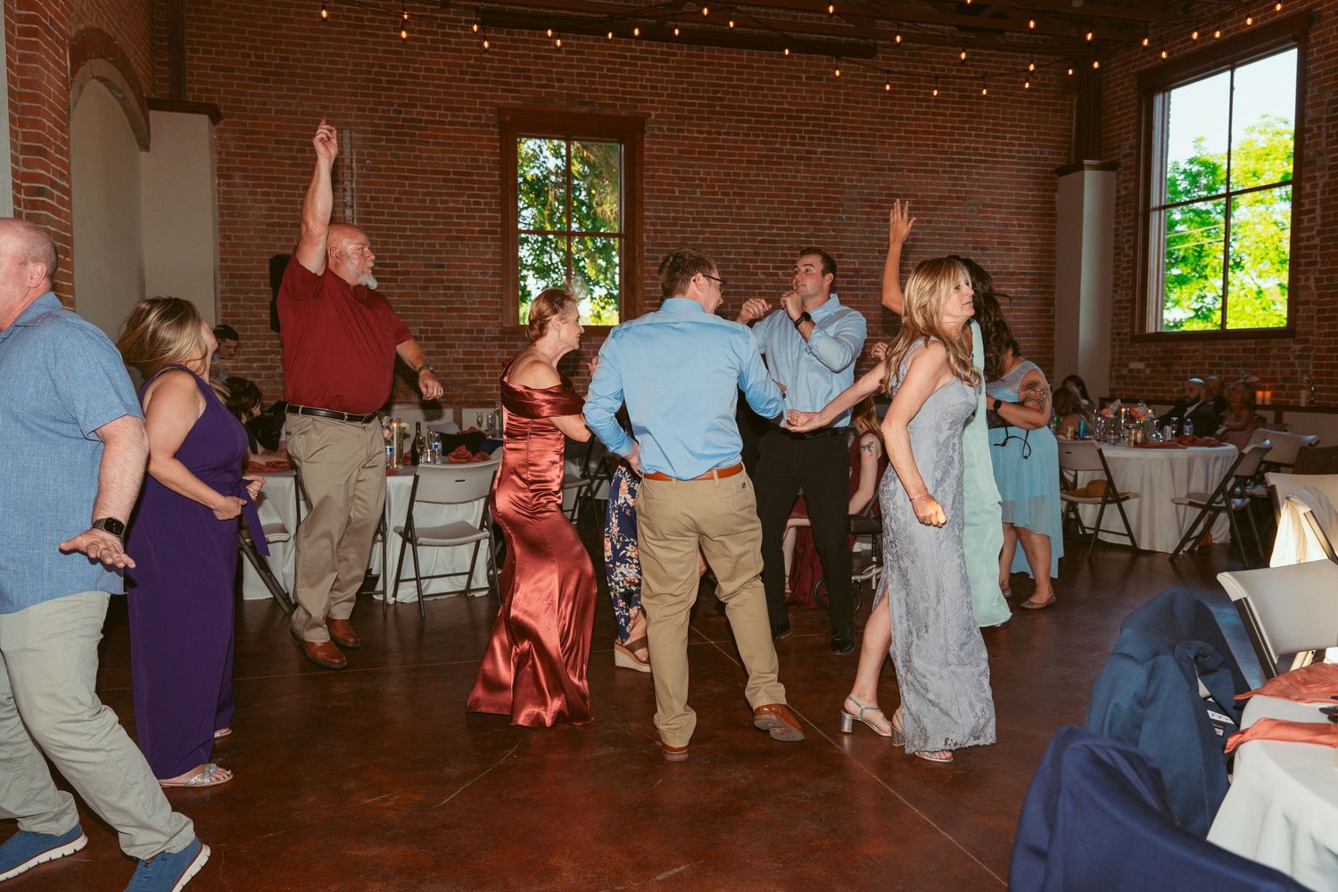 Wedding party members dancing in a line formation inside a rustic reception venue with exposed brick walls.