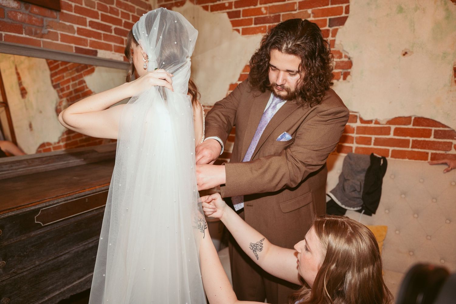 Wedding party members help adjust a bride's veil in a rustic brick-walled room.