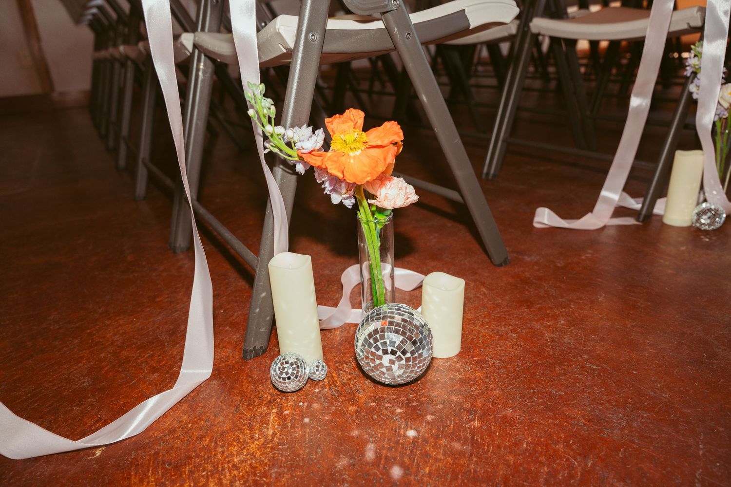 Orange flowers in small vase with white ribbons and candles on rustic floor.