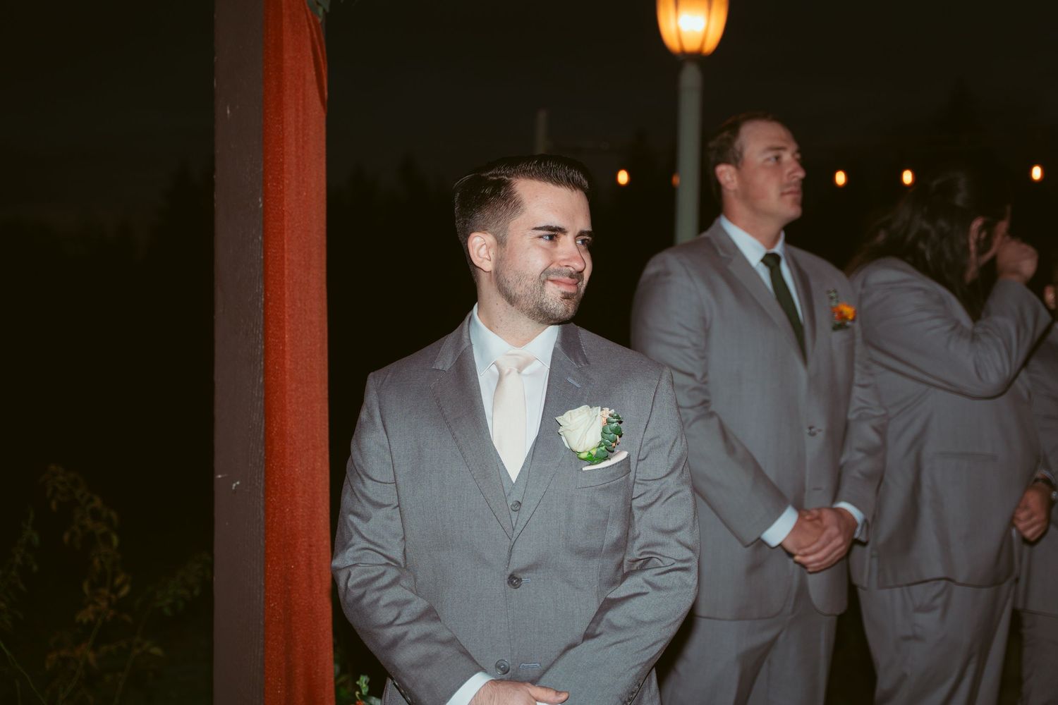 Groomsmen in gray suits stand together under amber lighting at an evening wedding reception.