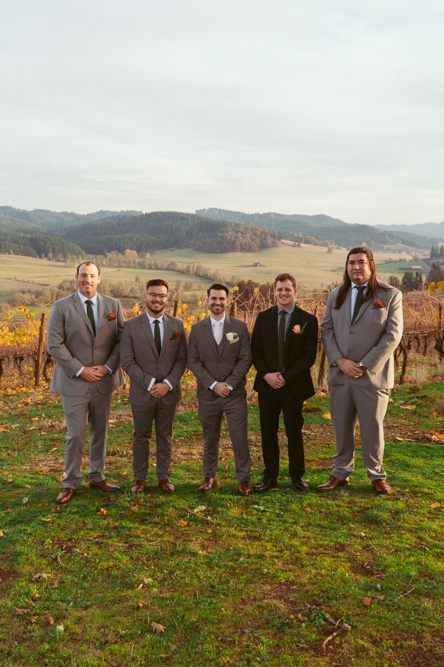 Wedding party members in coordinated grey suits pose for formal photo in front of autumn vineyard.