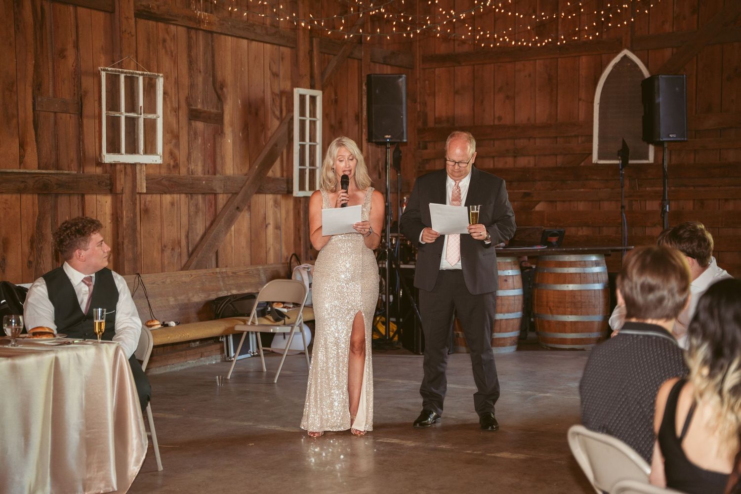 Wedding speeches in rustic barn venue with wine barrels and string light decorations.