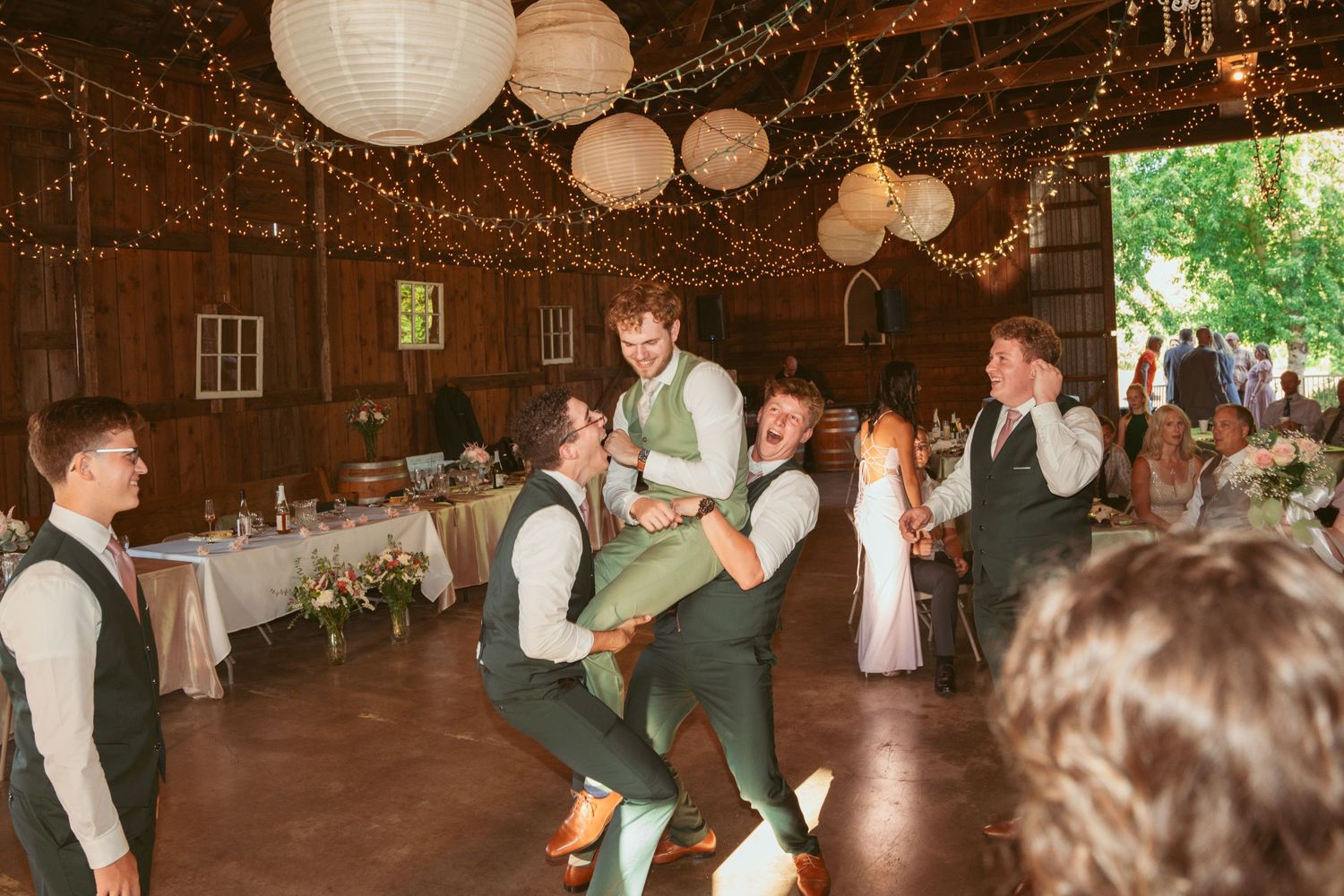 Guests participate in group dancing at a rustic barn wedding reception under warm lighting.
