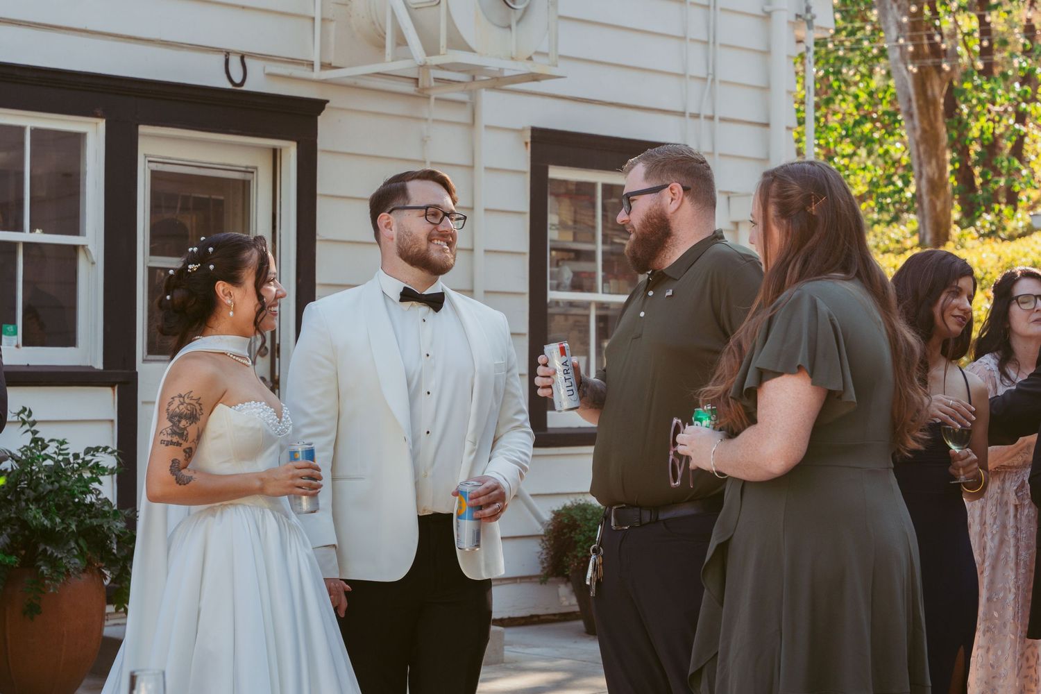 Wedding party members gather outside a traditional white house during a summer celebration.