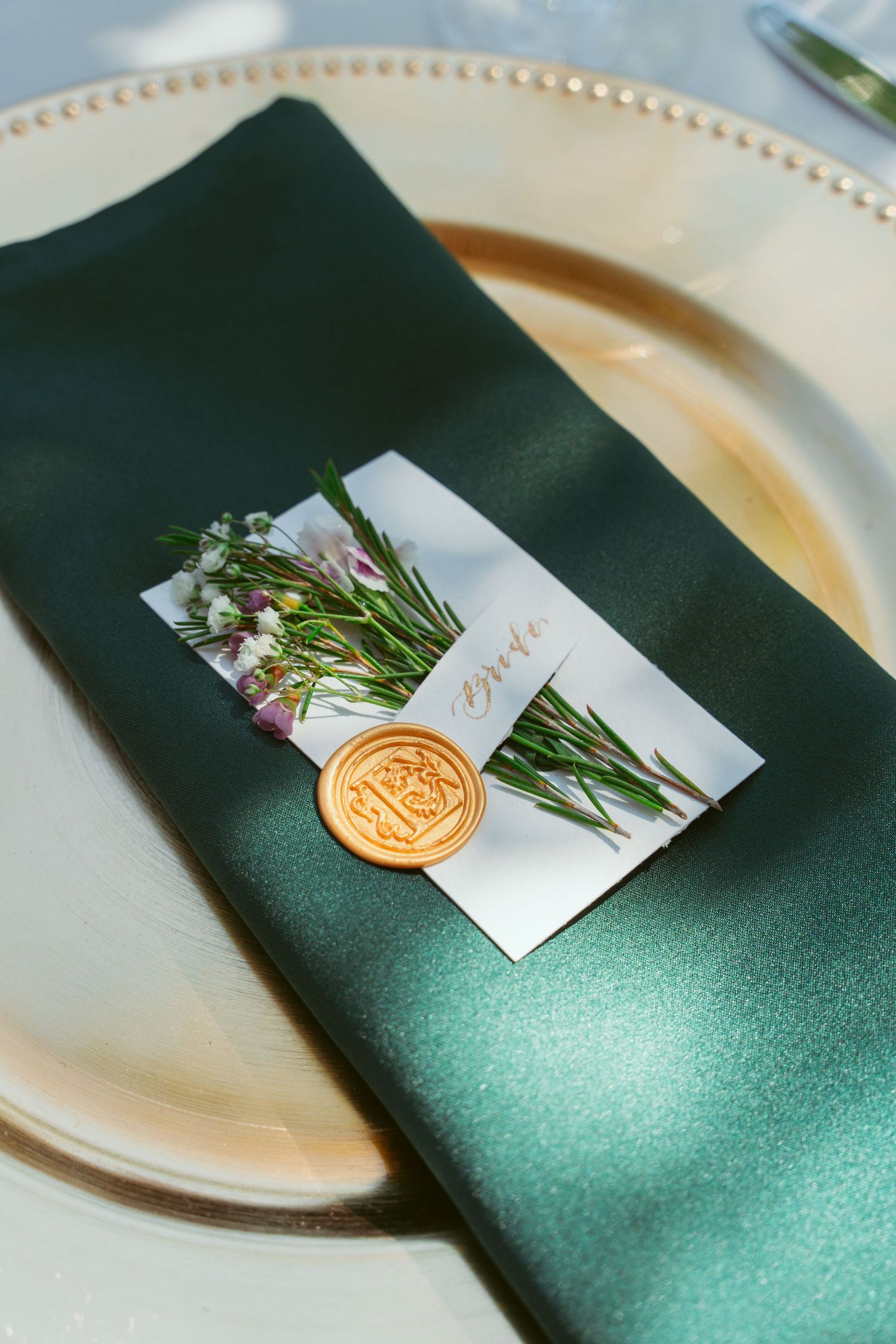 Elegant place setting with dark green napkin and gold wax seal accented with fresh herbs and floral details.