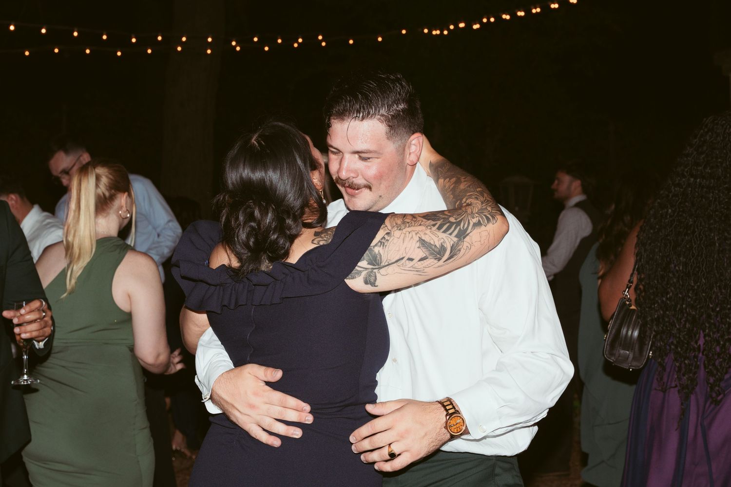 Couples slow dancing at an evening outdoor wedding reception with string lights overhead.