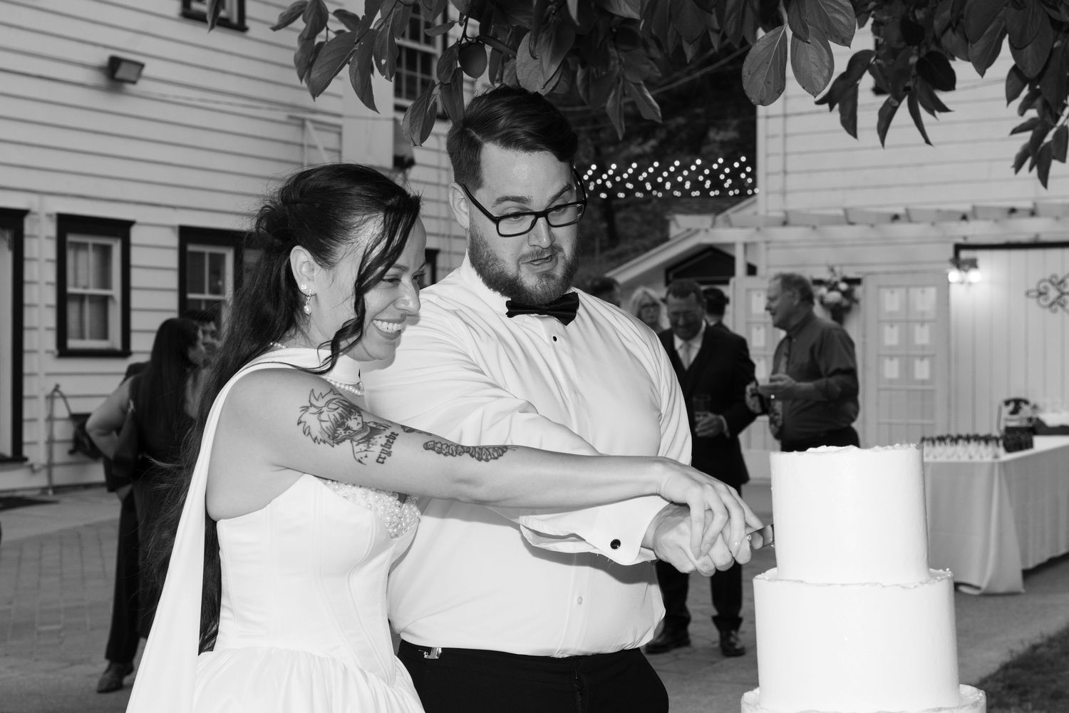 A couple laughs together while serving their wedding cake at an outdoor celebration.