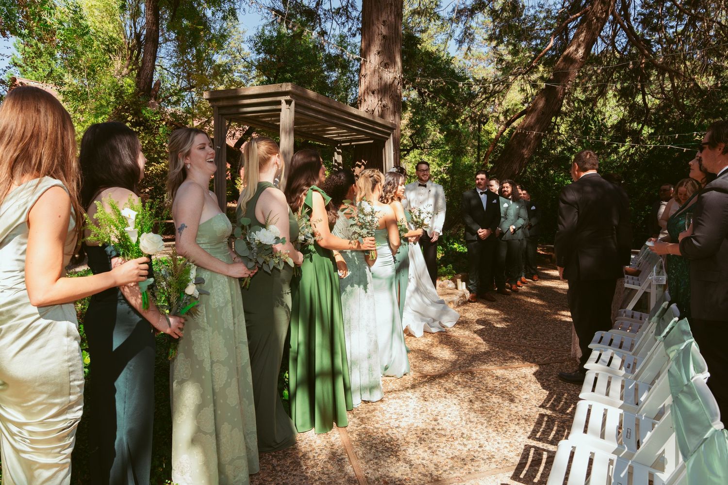 Wedding party in pastel dresses stands in line during outdoor garden ceremony.