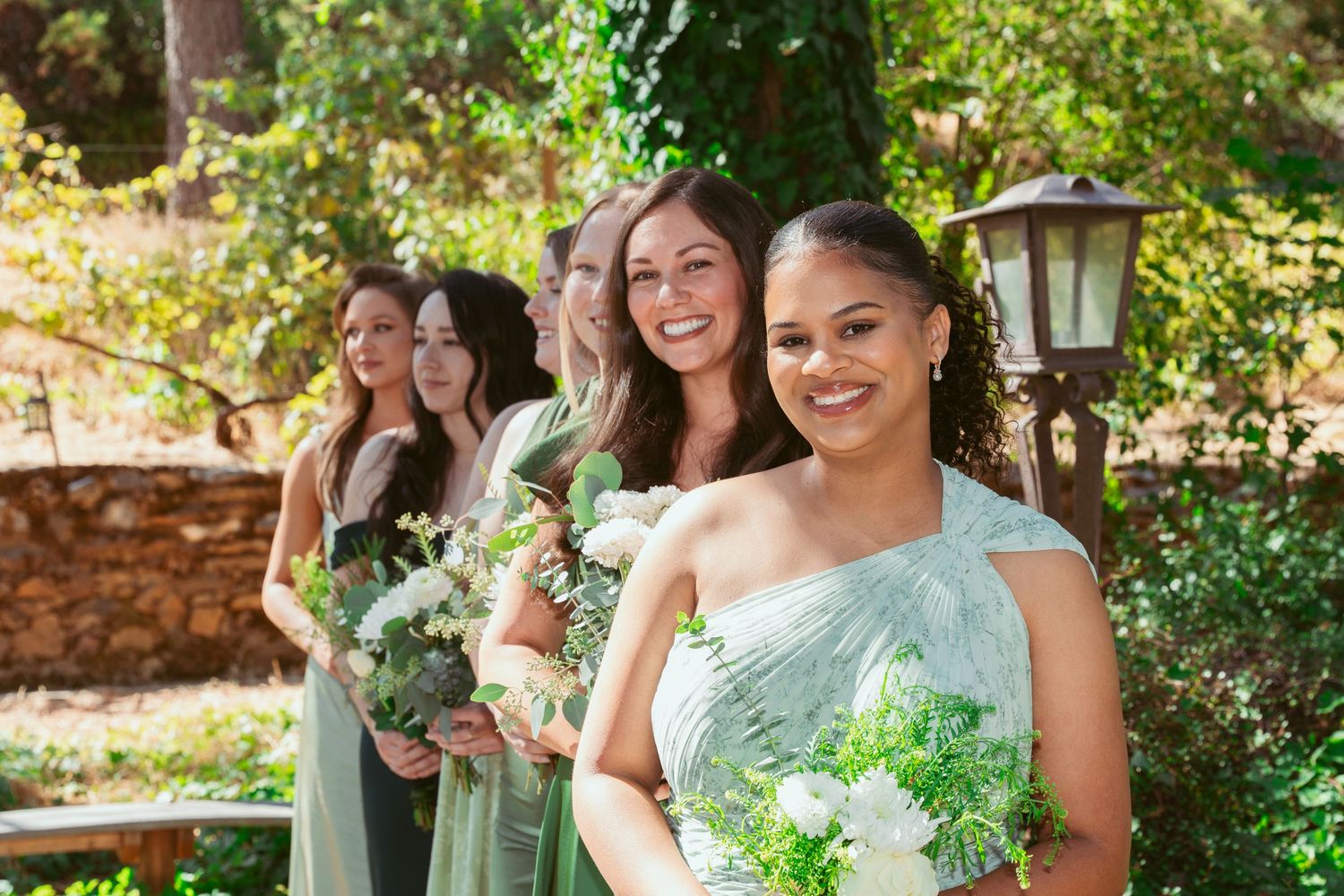 Bridesmaids in sage green dresses hold white bouquets while standing outdoors in a garden setting.