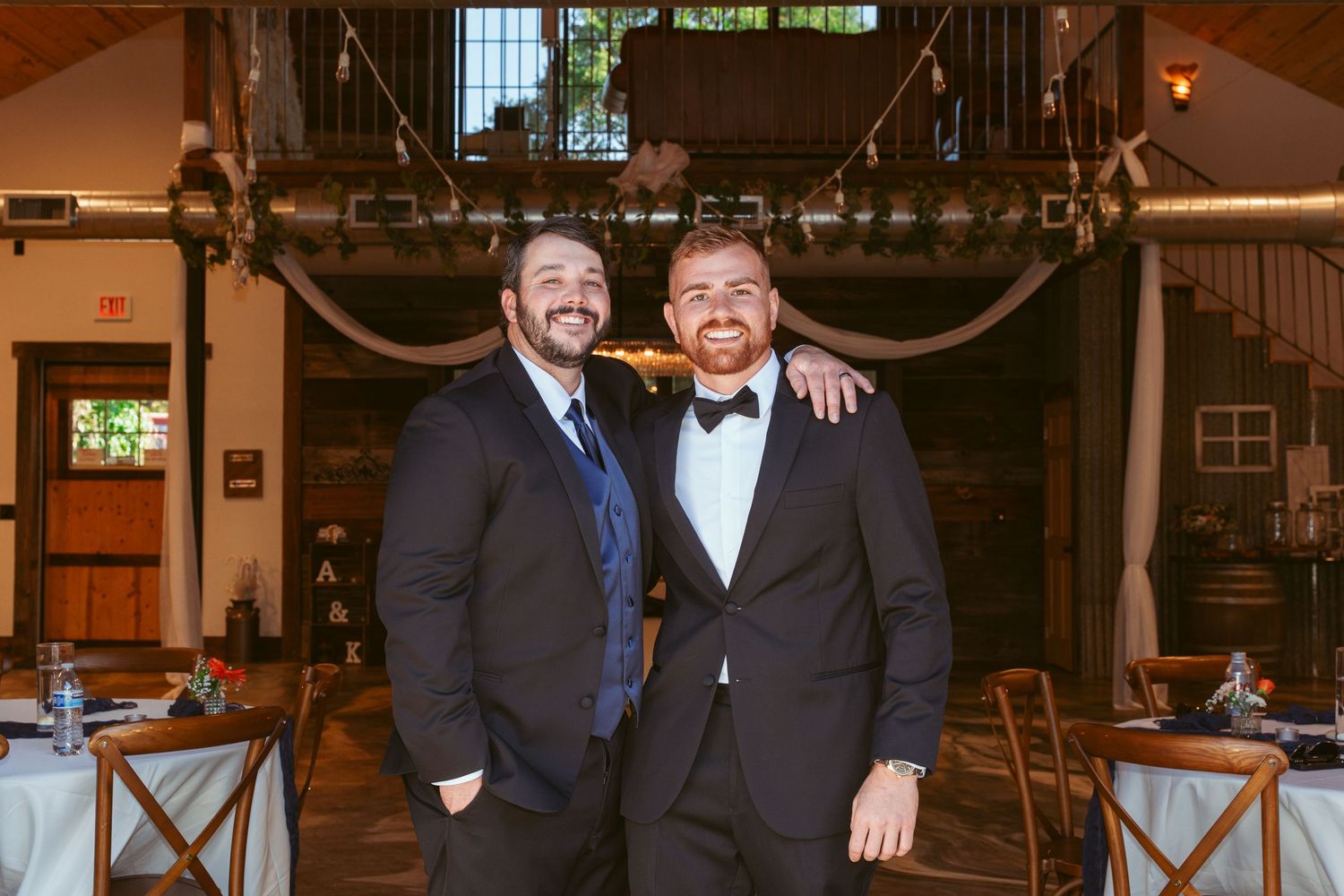 Two men in dark suits posing together at an industrial-style wedding venue.