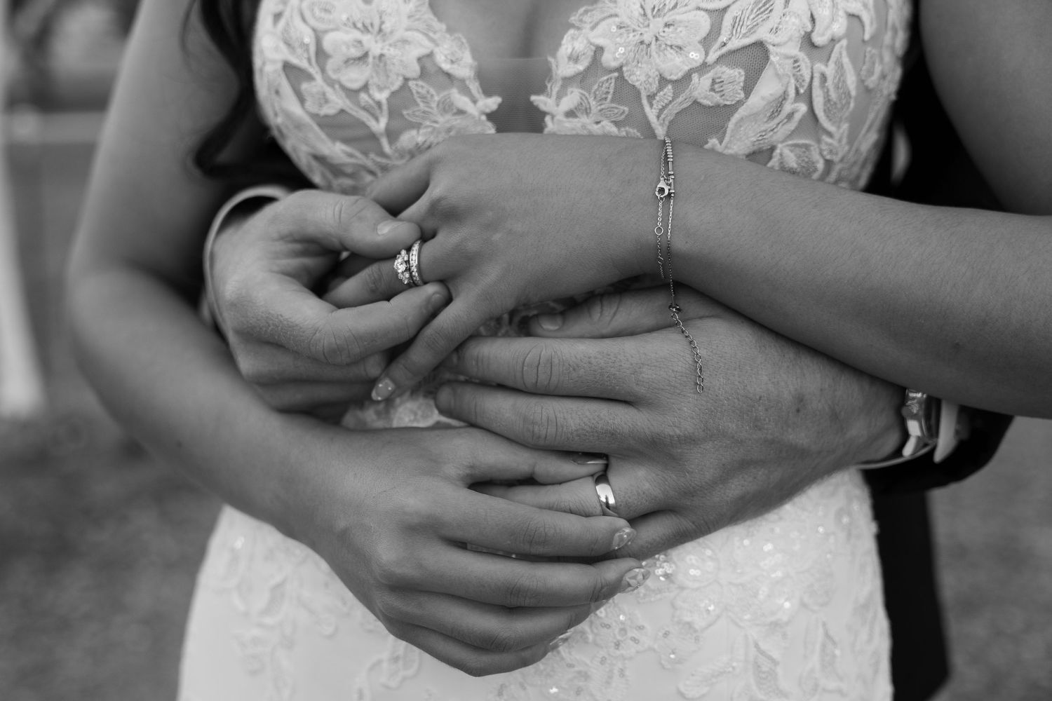 Close-up black and white detail of hands with wedding rings and lace dress bodice.