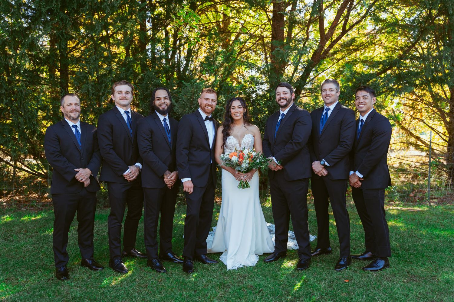 Wedding party in navy suits and white gown pose together on grass with trees in background.