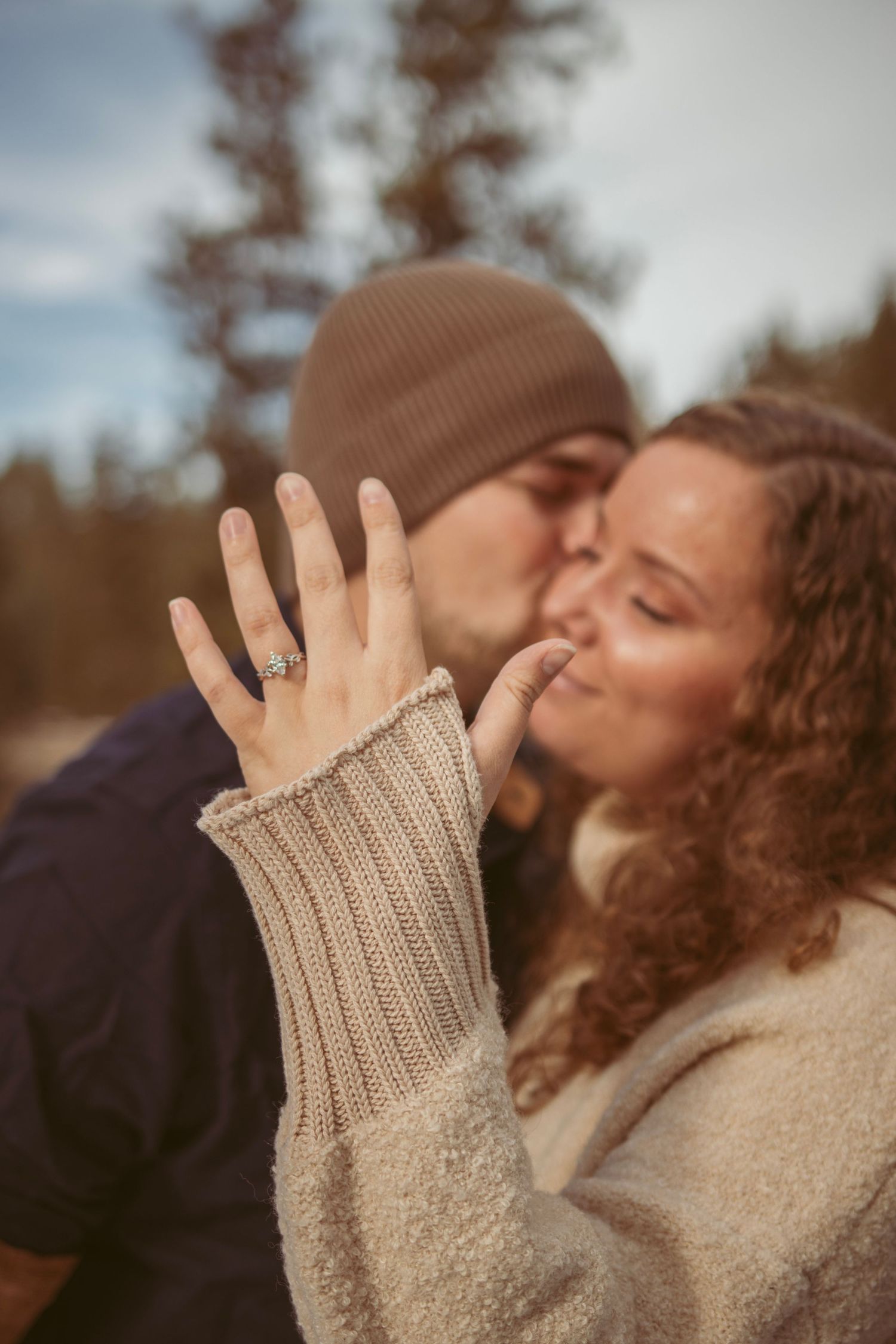 Romantic couple sharing a tender kiss outdoors while showing off an engagement ring in winter.