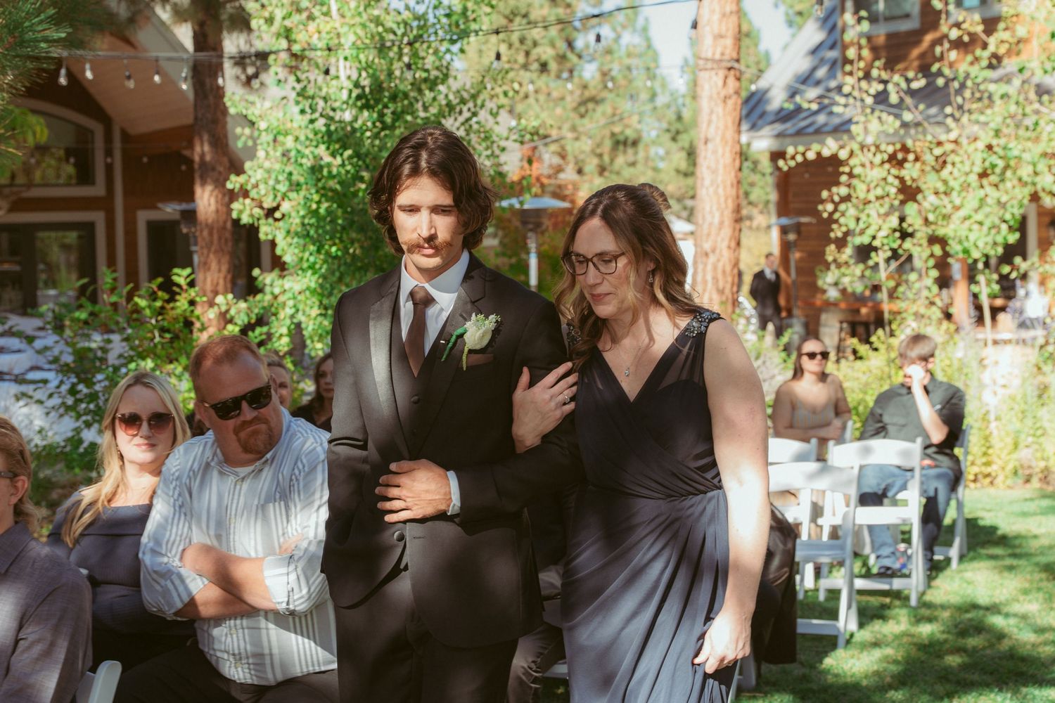 Wedding party members process down an aisle at an outdoor ceremony with pine trees overhead.