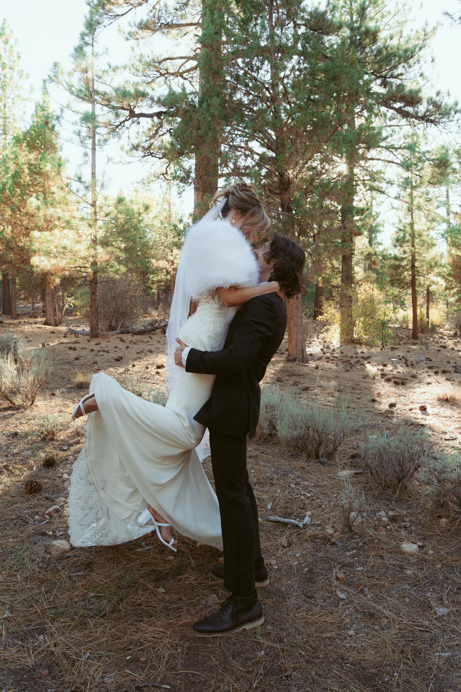 Romantic wedding couple embracing in a pine forest setting with natural lighting and fallen leaves.