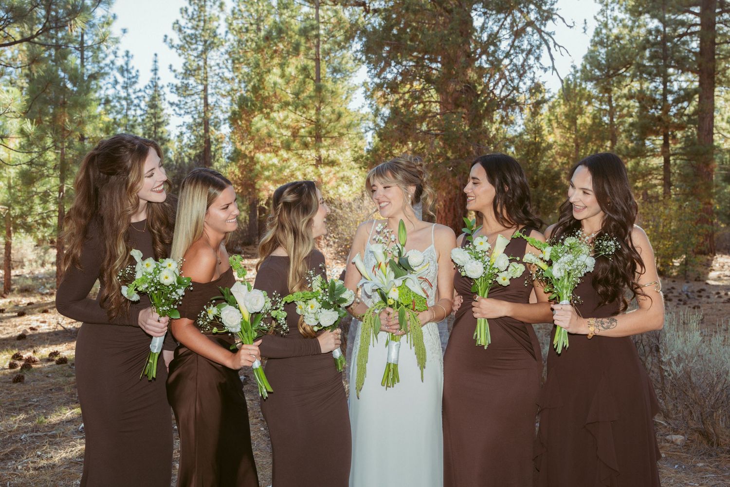 Bridesmaids in brown dresses hold white bouquets while posing in natural forest setting.