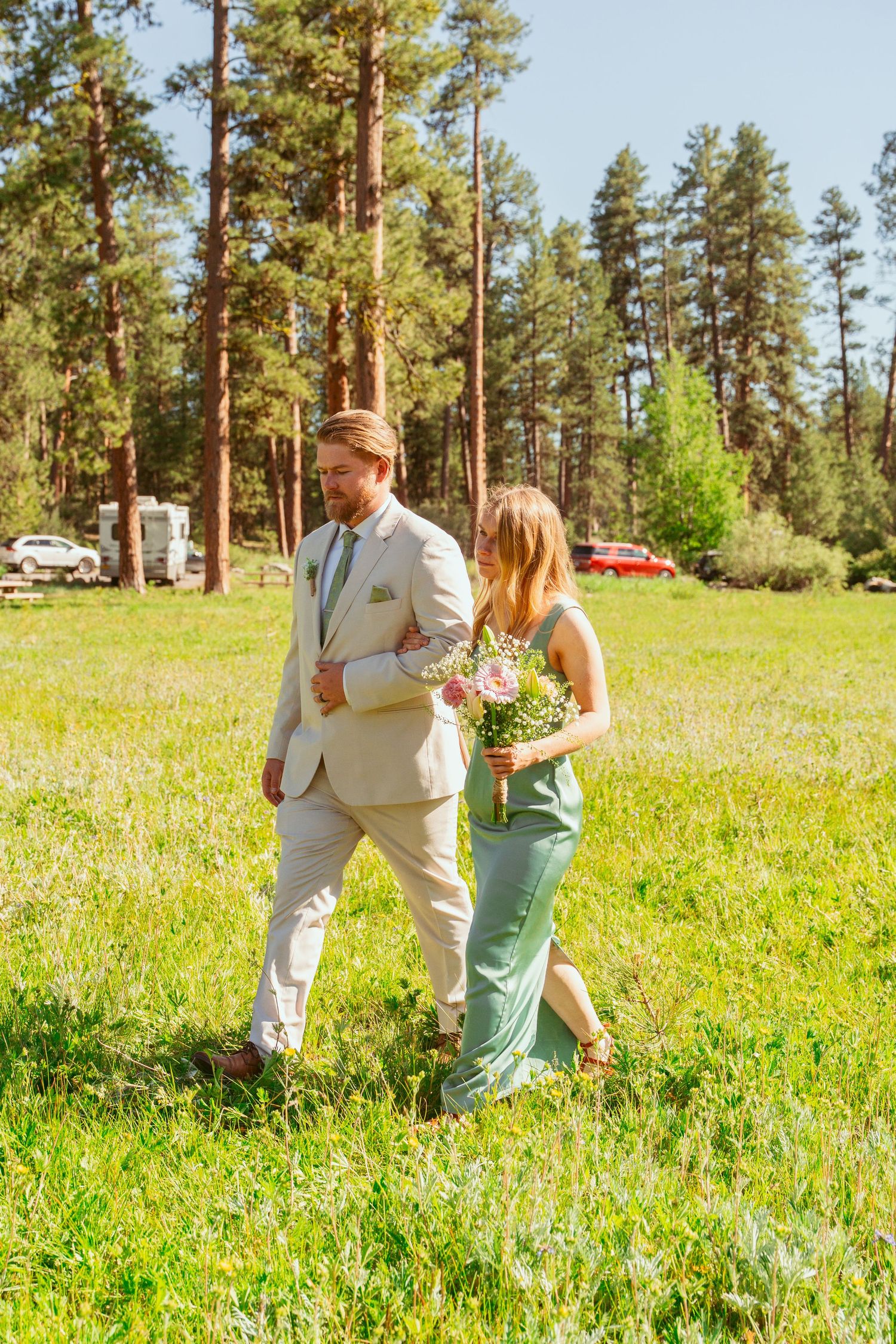 Wedding attendants in formal attire walking through a wildflower meadow with pine trees.