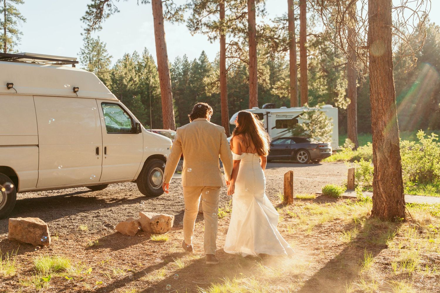 Sun-lit forest path with parked vehicles and wedding attendees in light-colored attire.
