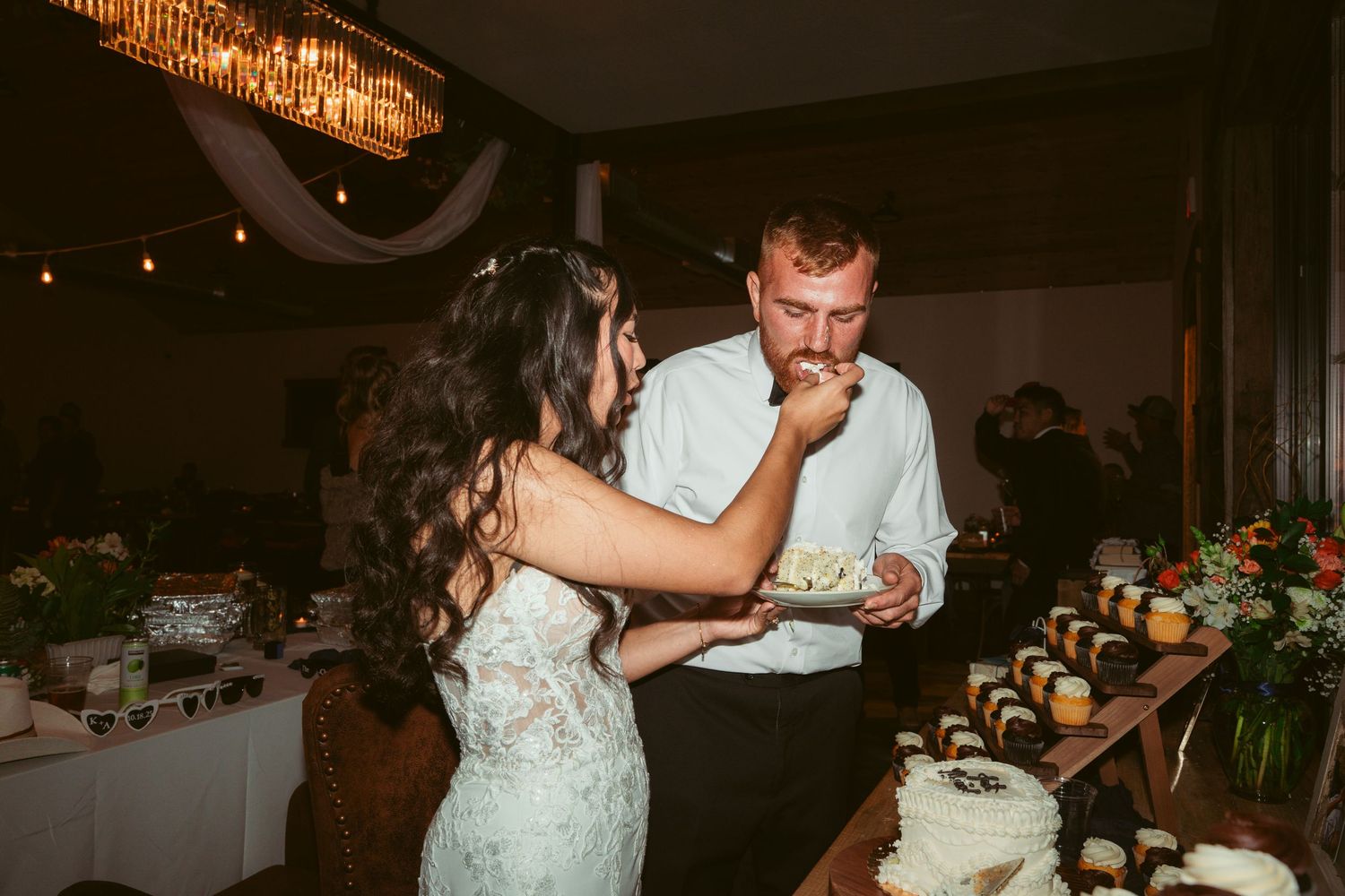 Evening wedding reception features couple participating in cake cutting ceremony under ambient lighting.