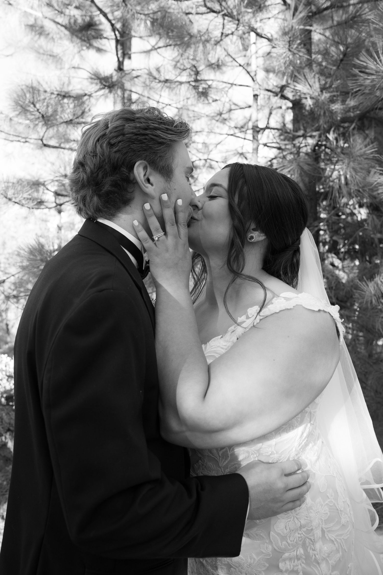 A romantic black and white wedding photo of a couple sharing their first kiss in front of pine trees.