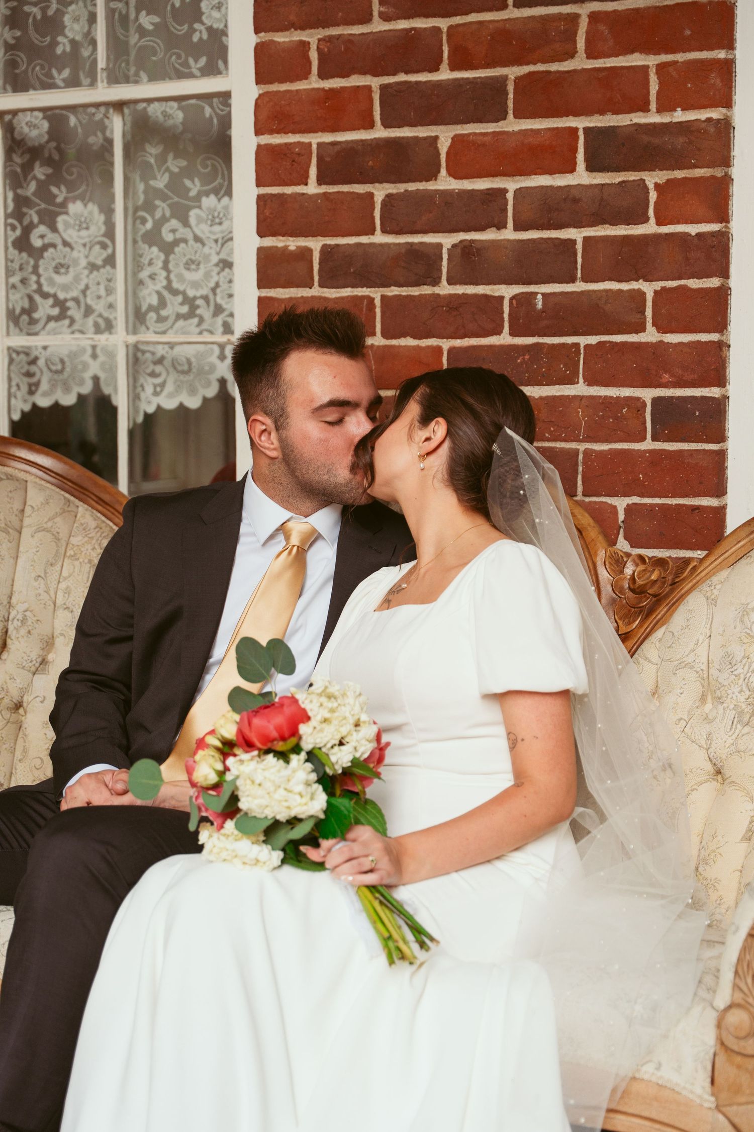 An intimate moment between newlyweds sharing a kiss on an antique sofa with a floral bouquet.