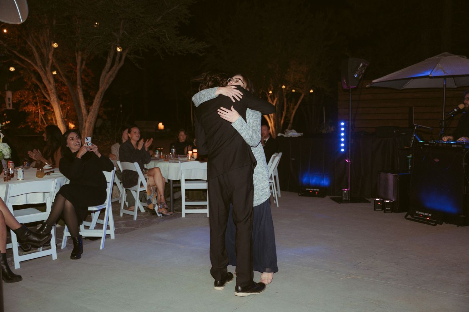 A couple embraces while slow dancing at an outdoor reception with guests seated nearby.