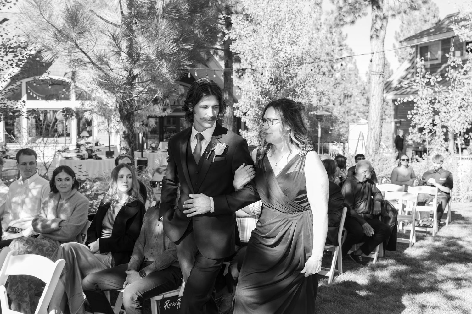 Black and white photo of wedding party members walking down an outdoor aisle with guests seated.