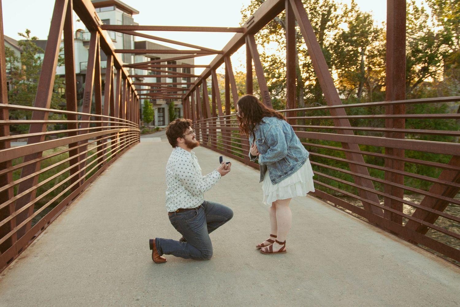 A surprise proposal taking place on a wooden pedestrian bridge at dusk.