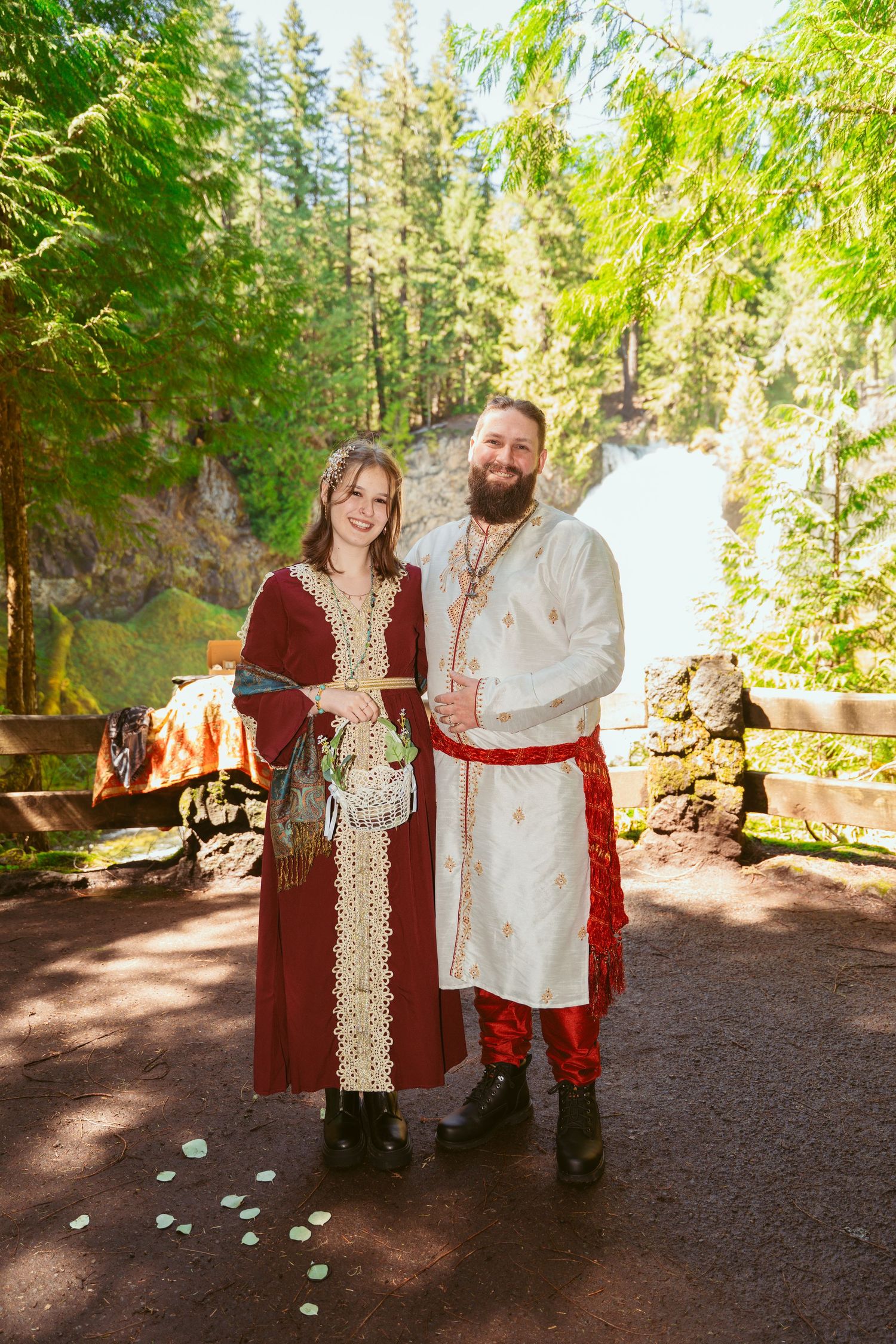 A medieval-themed wedding party stands together on a dirt path surrounded by tall evergreen trees.