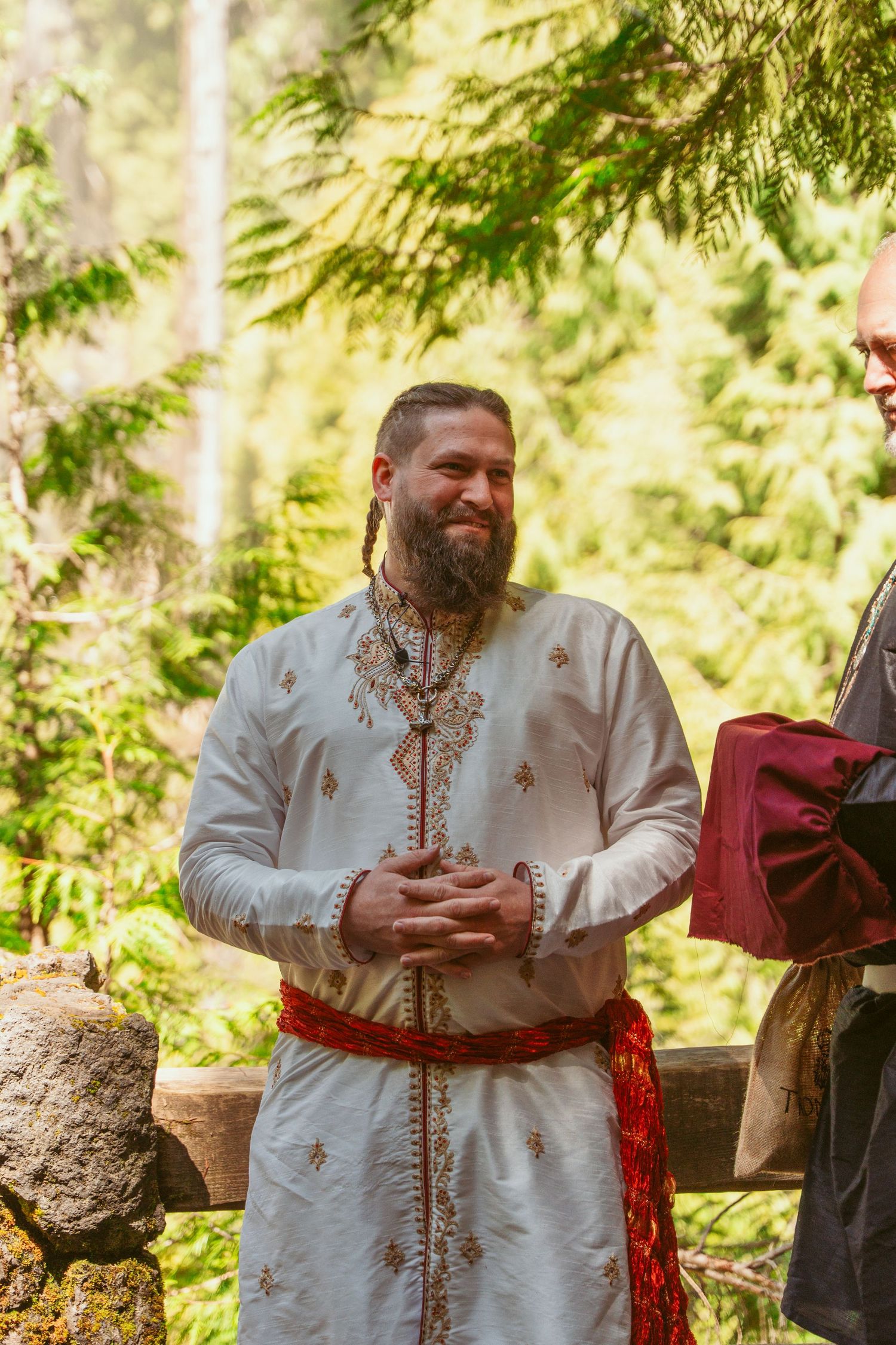 Medieval-style ceremony in a forest setting with a person in white embroidered tunic and red sash.