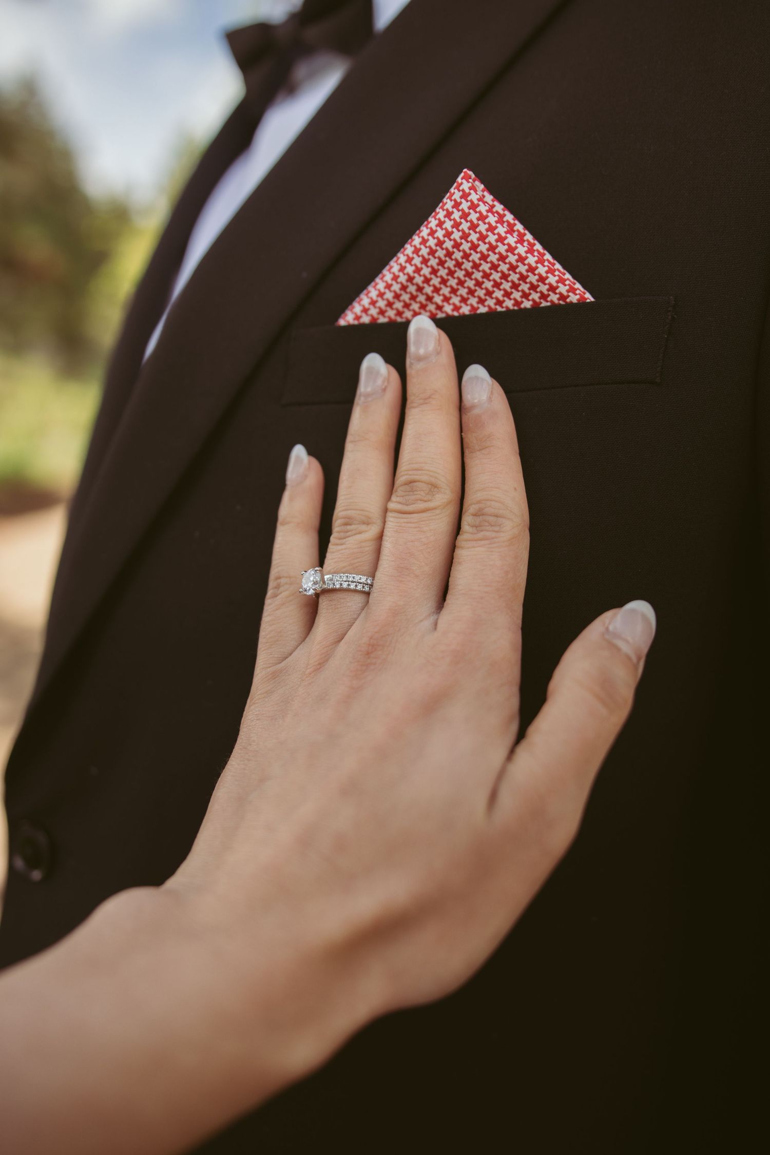 Manicured hand with wedding ring touching black suit jacket with red checkered pocket square.