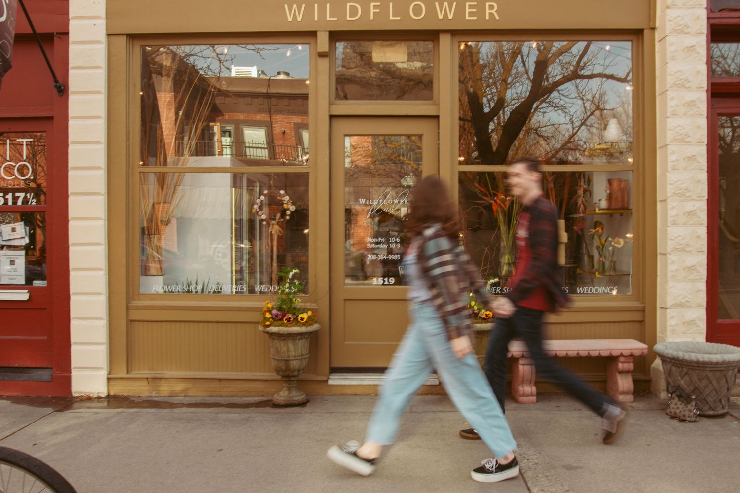 Motion-blurred pedestrians pass by the entrance of a Wildflower shop with vintage-style storefront.