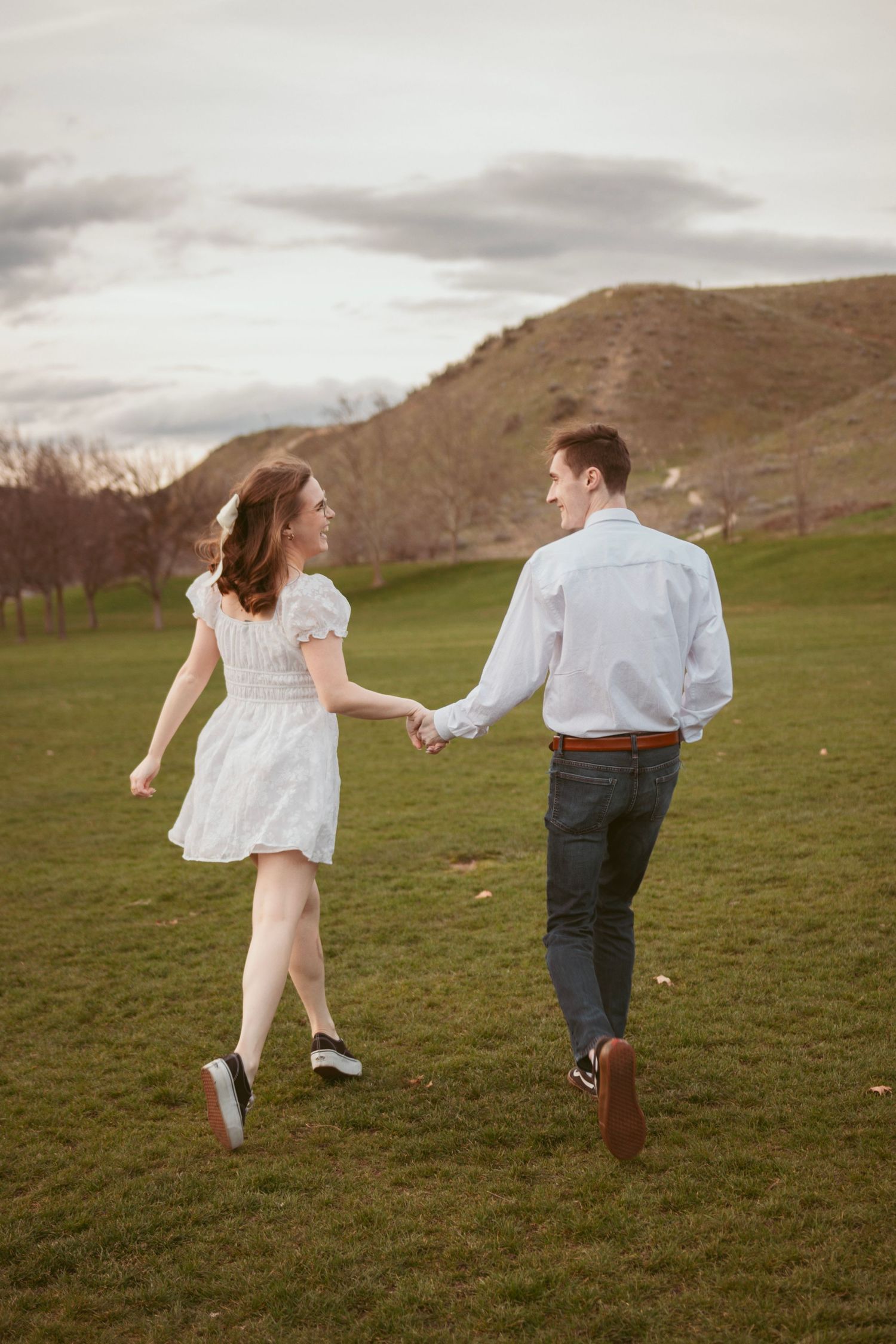 Two people skip playfully through a grassy meadow with mountains visible in the distance.