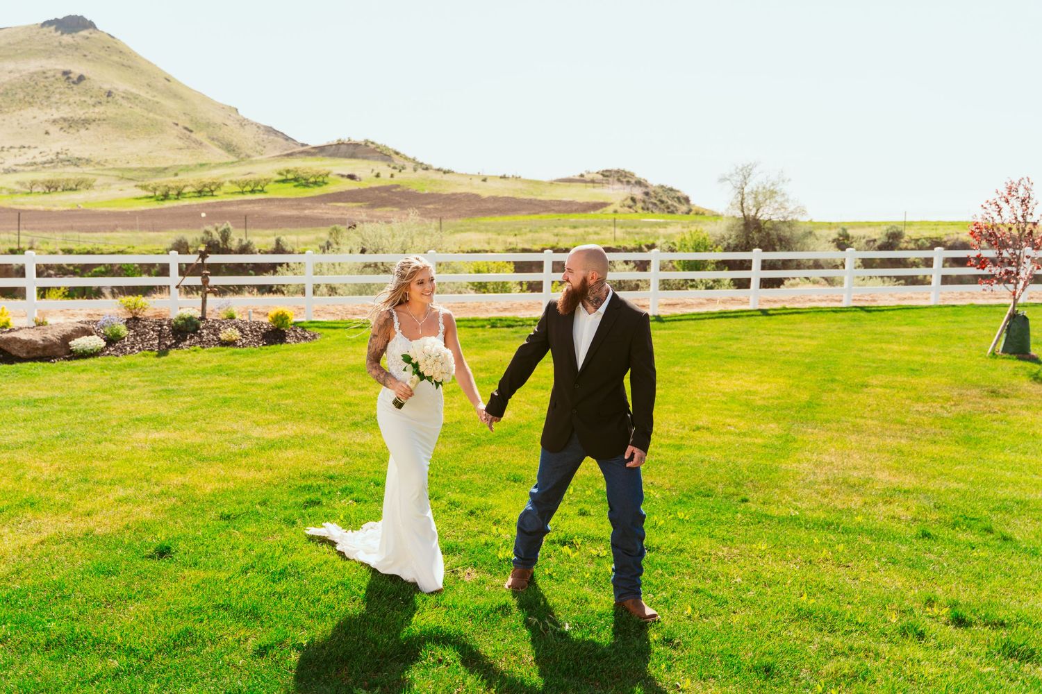Newlyweds walking hand in hand on green grass with mountains in background.
