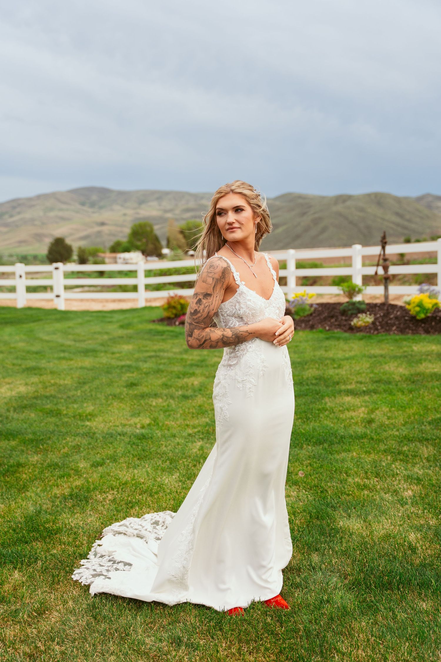 Bride in fitted lace gown with red shoes poses in front of mountain landscape and white fence.