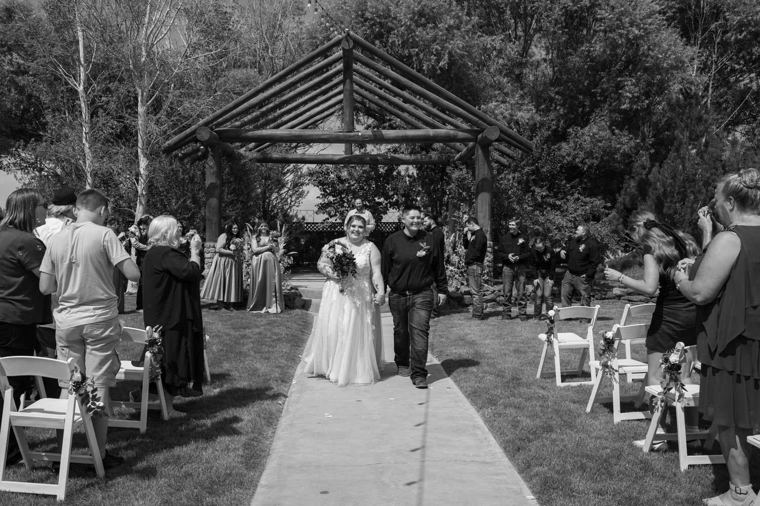 Black and white photo of wedding guests seated outdoors at a garden ceremony with trees in the background.