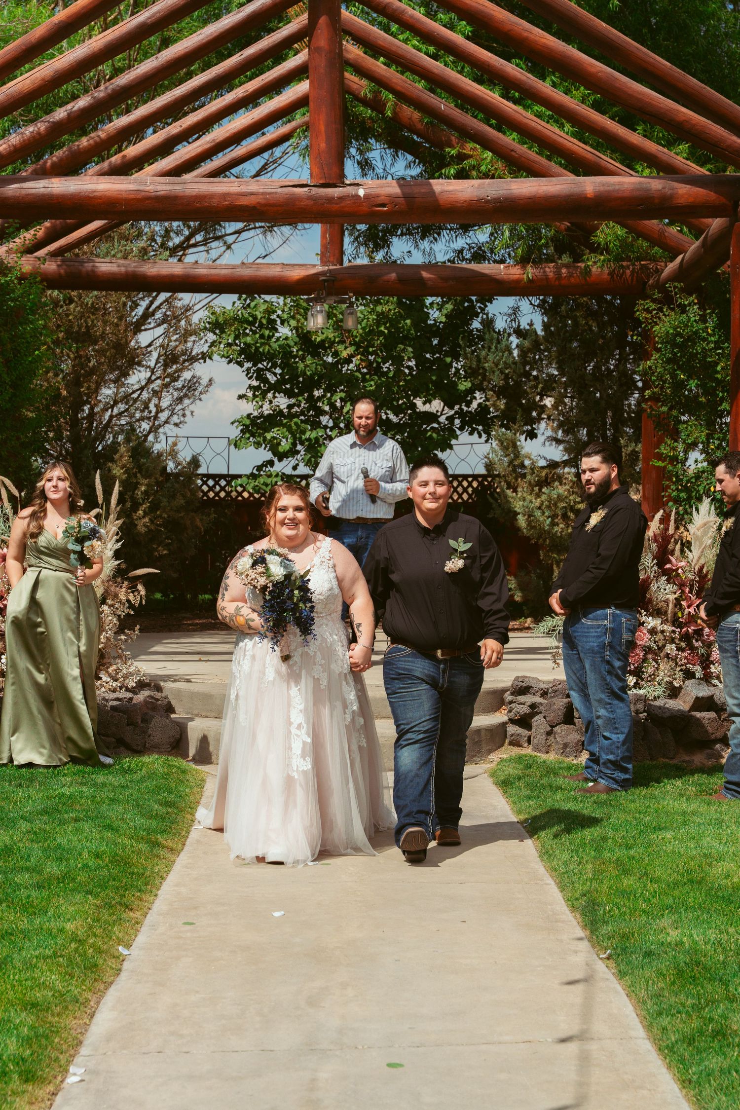 Wedding guests and participants gather under a wooden pergola walkway during an outdoor ceremony.