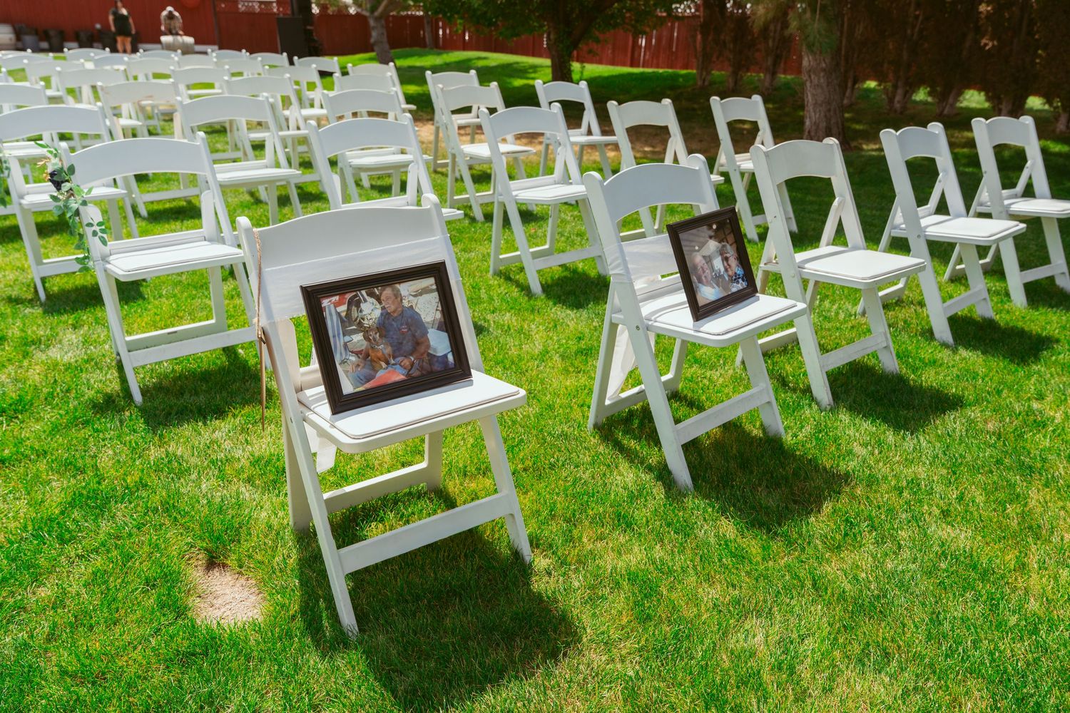 White folding chairs with framed photos displayed on seats arranged on green grass for a memorial or tribute ceremony.