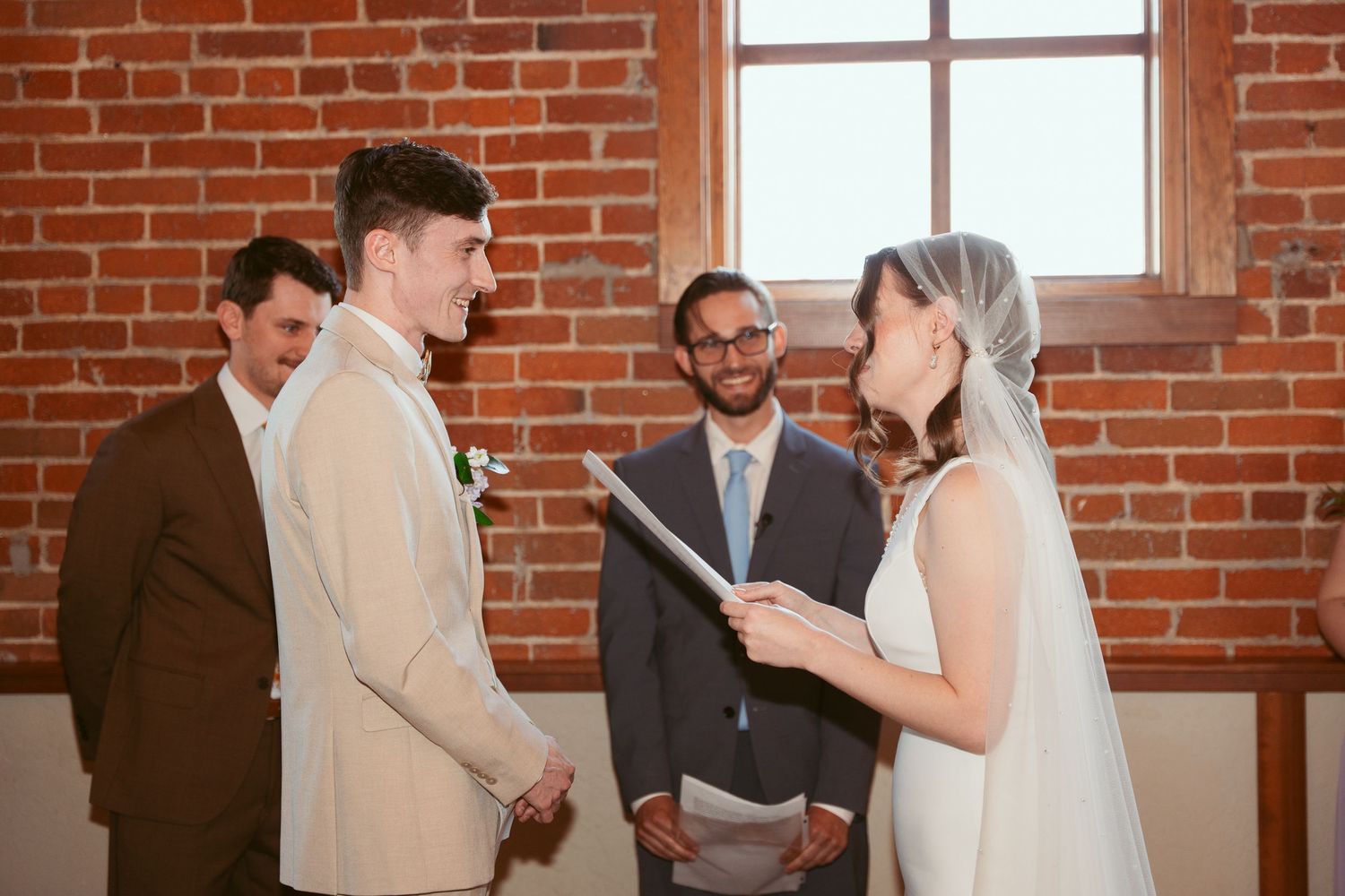 Wedding ceremony taking place in a bright brick-walled venue with large windows.