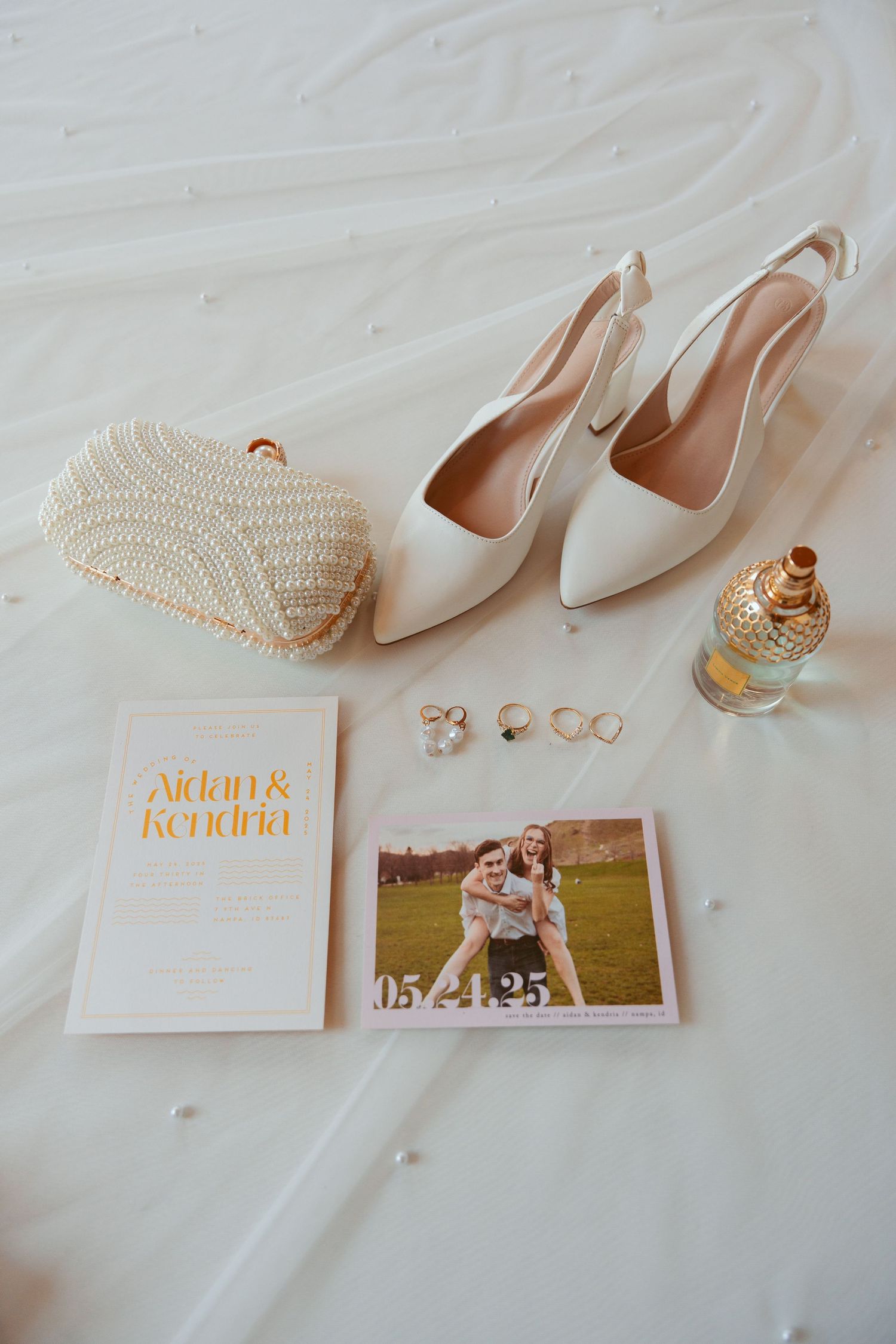 Wedding flat lay with white shoes, pearl clutch, rings, invitation and engagement photo on white background.