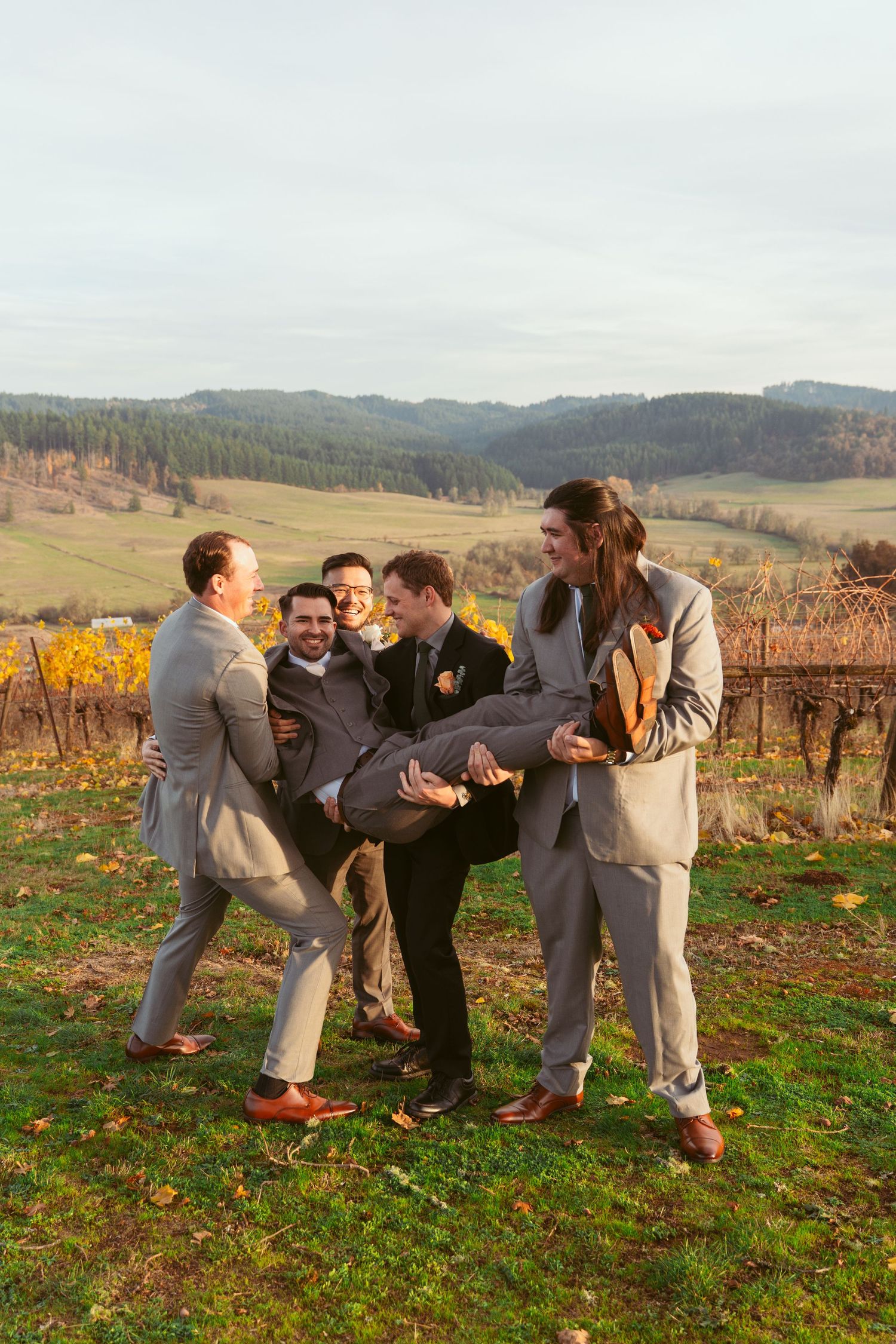 Groomsmen in grey suits having fun and lifting someone during outdoor wedding photos.