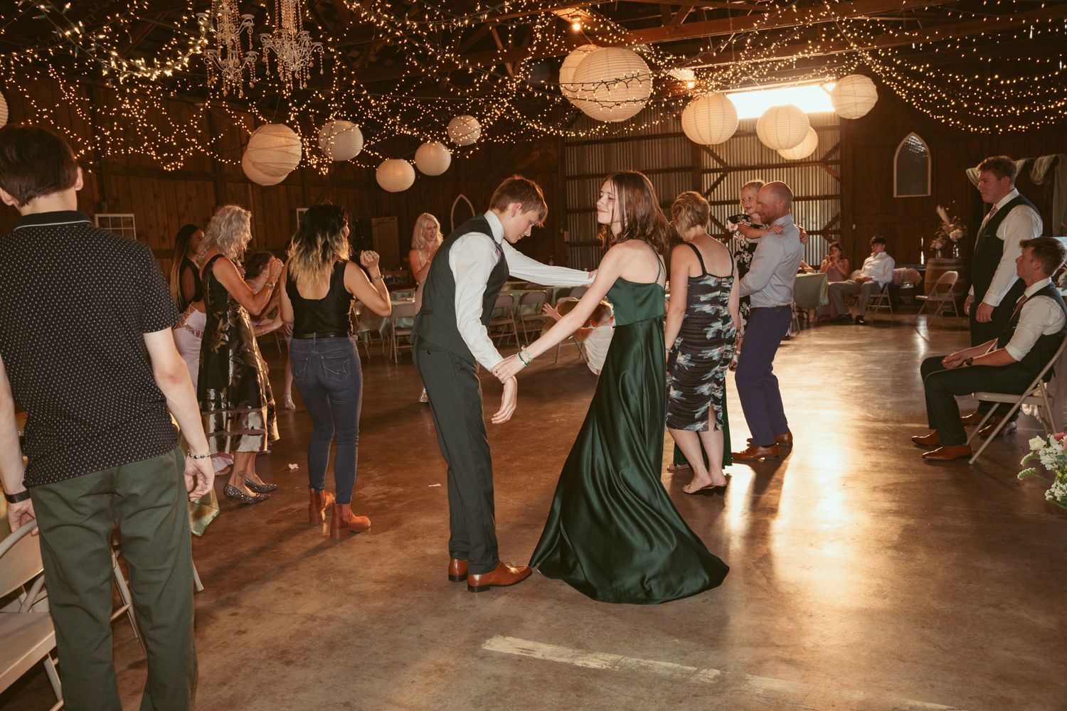 Couples dancing together at a rustic barn reception with twinkle lights and paper lanterns overhead.